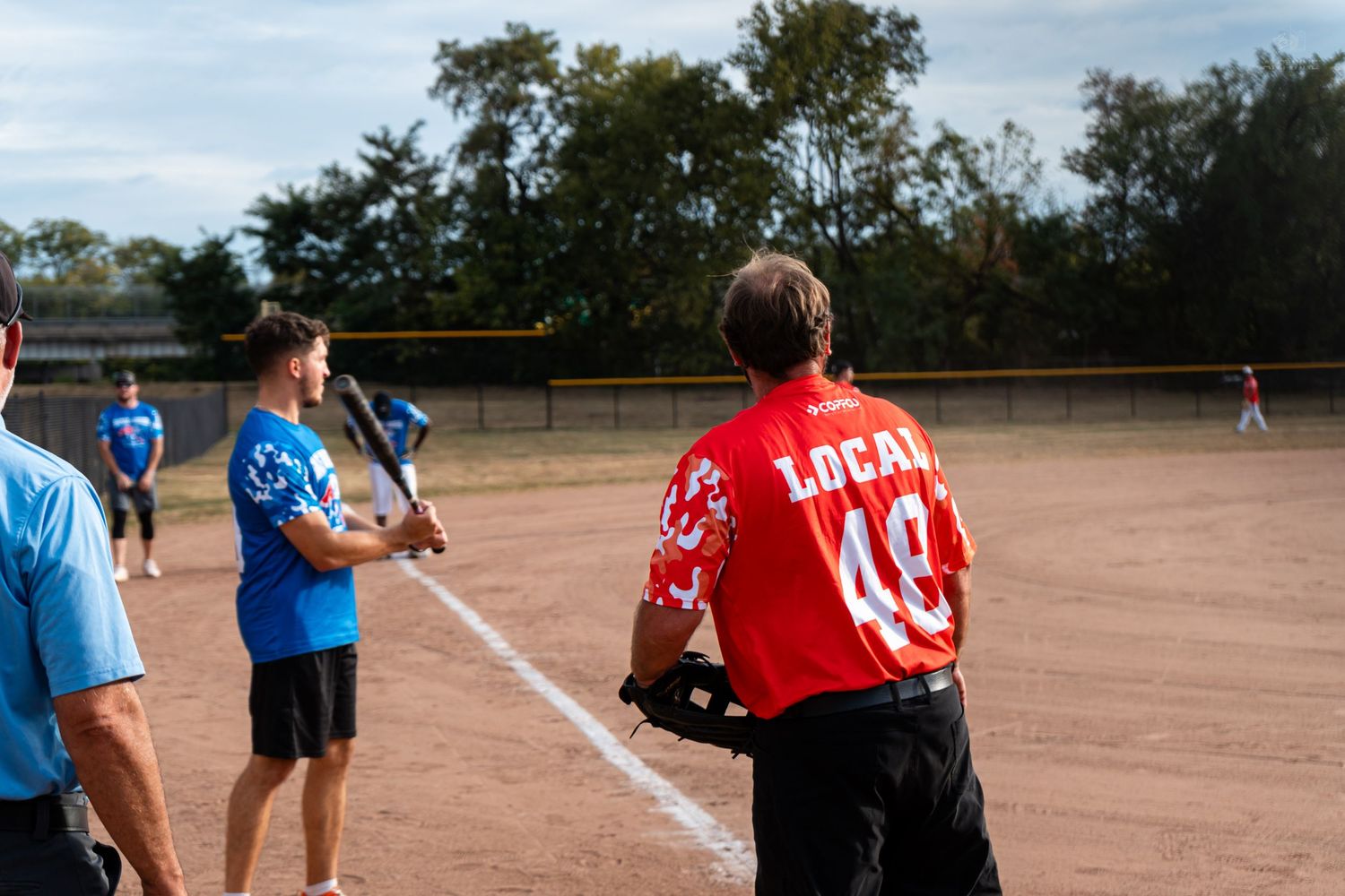 A player in a red Local 48 jersey watches from the sideline holding a bat during a softball game.