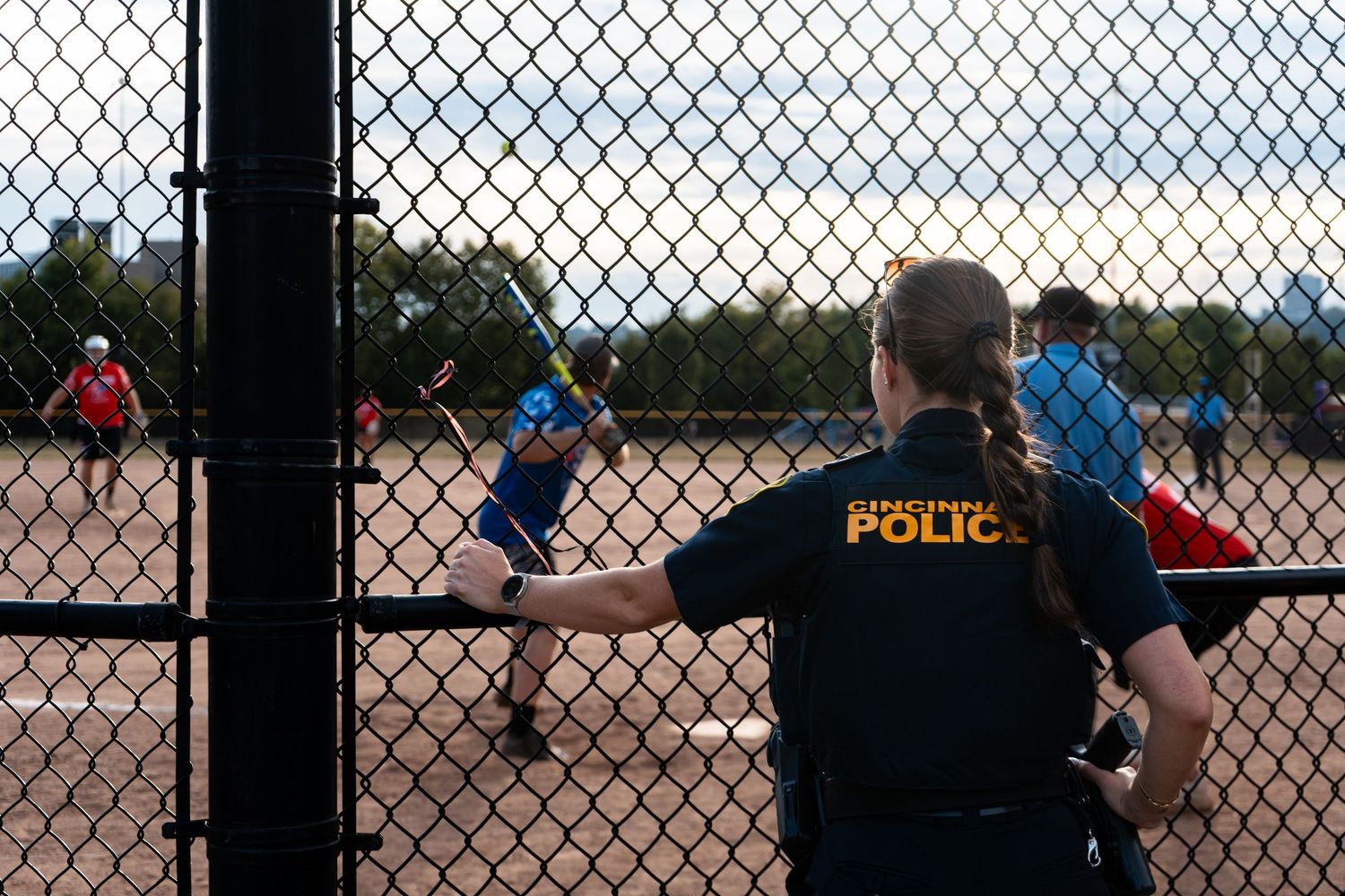 Police officer watches youth baseball game through chain-link fence at dusk on outdoor diamond.