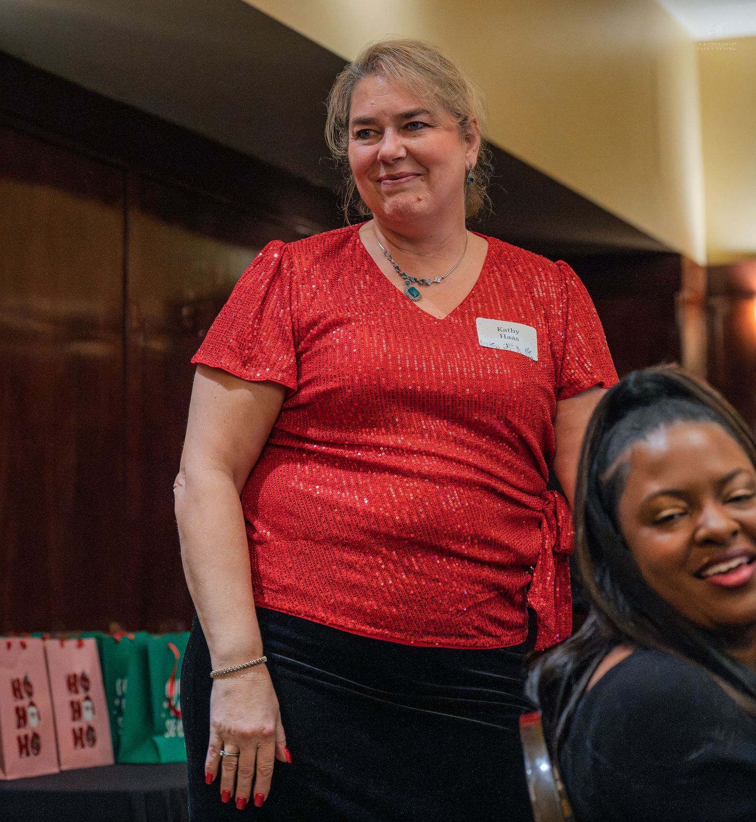 Woman in red sequin top wearing name tag smiles at indoor holiday party event.