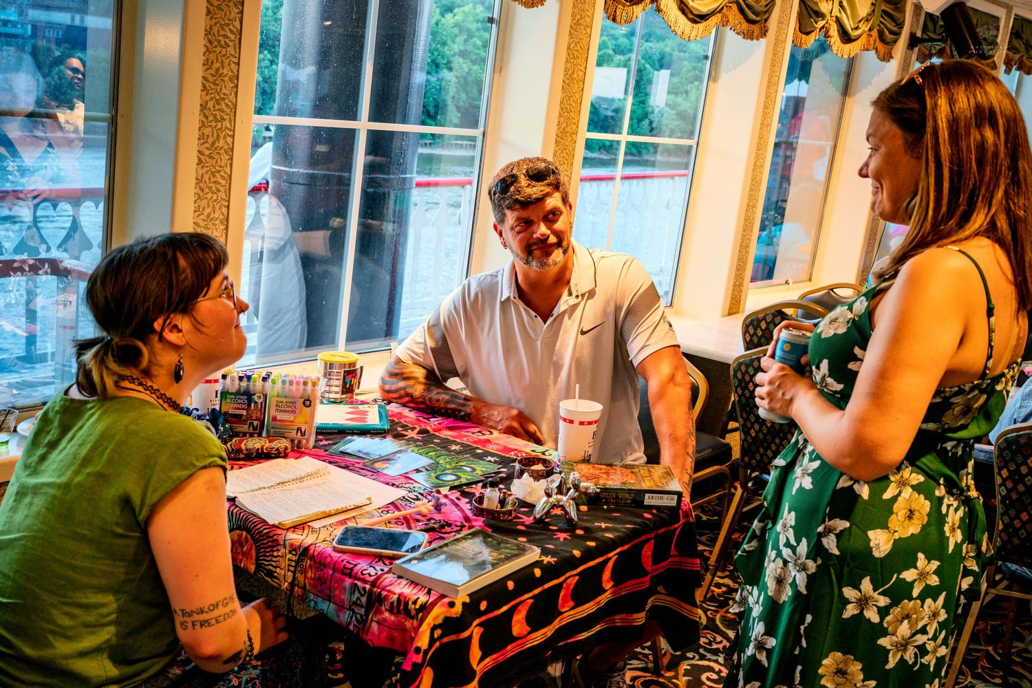 Three people socialize around a colorful table at an indoor gathering, with large windows in the background.