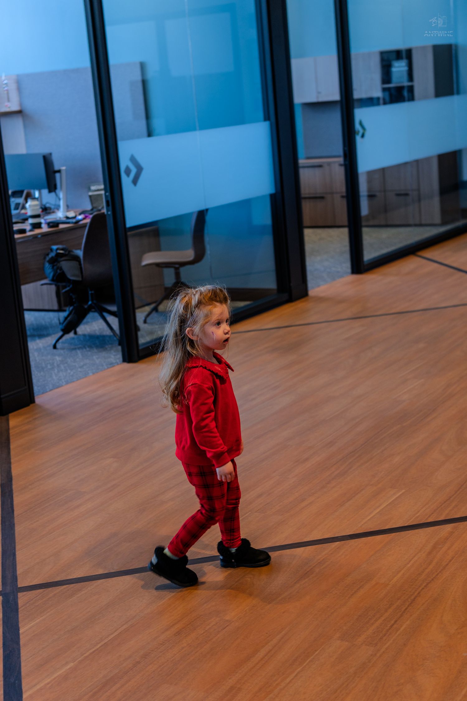 Young girl in red outfit walking through a modern office hallway with glass partition walls.