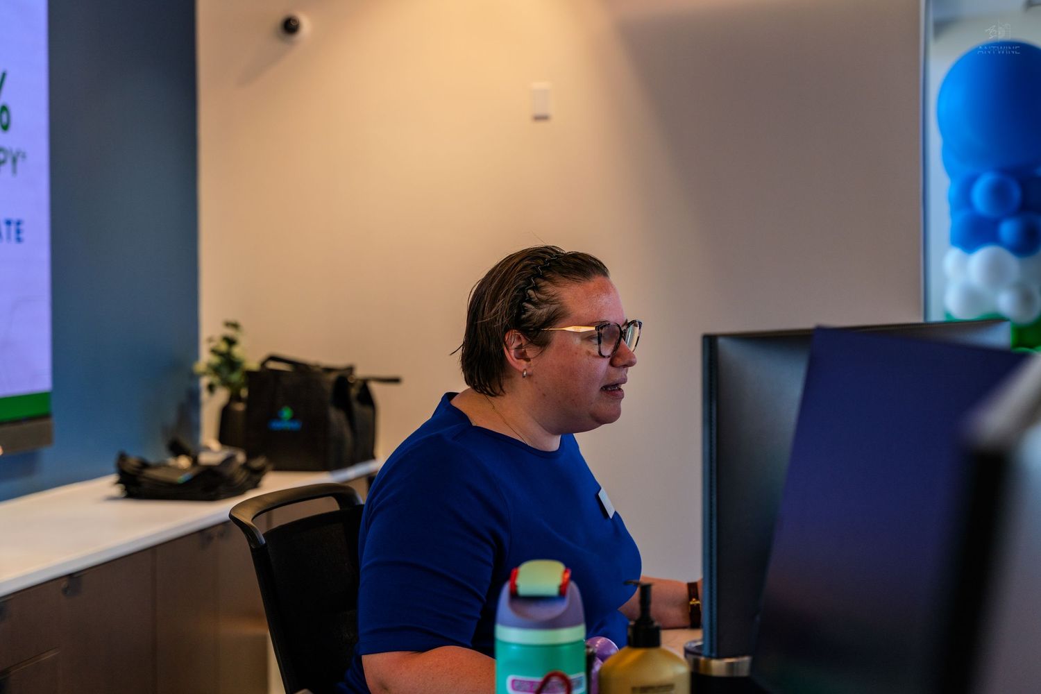 Professional working at dual monitors in modern office wearing blue shirt and glasses with decorative balloons in background.