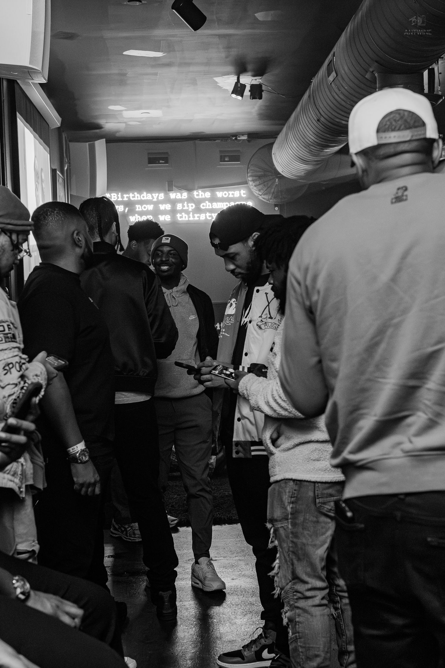 Black and white photo of a group of young men gathered indoors at a social event with neon text on the wall.