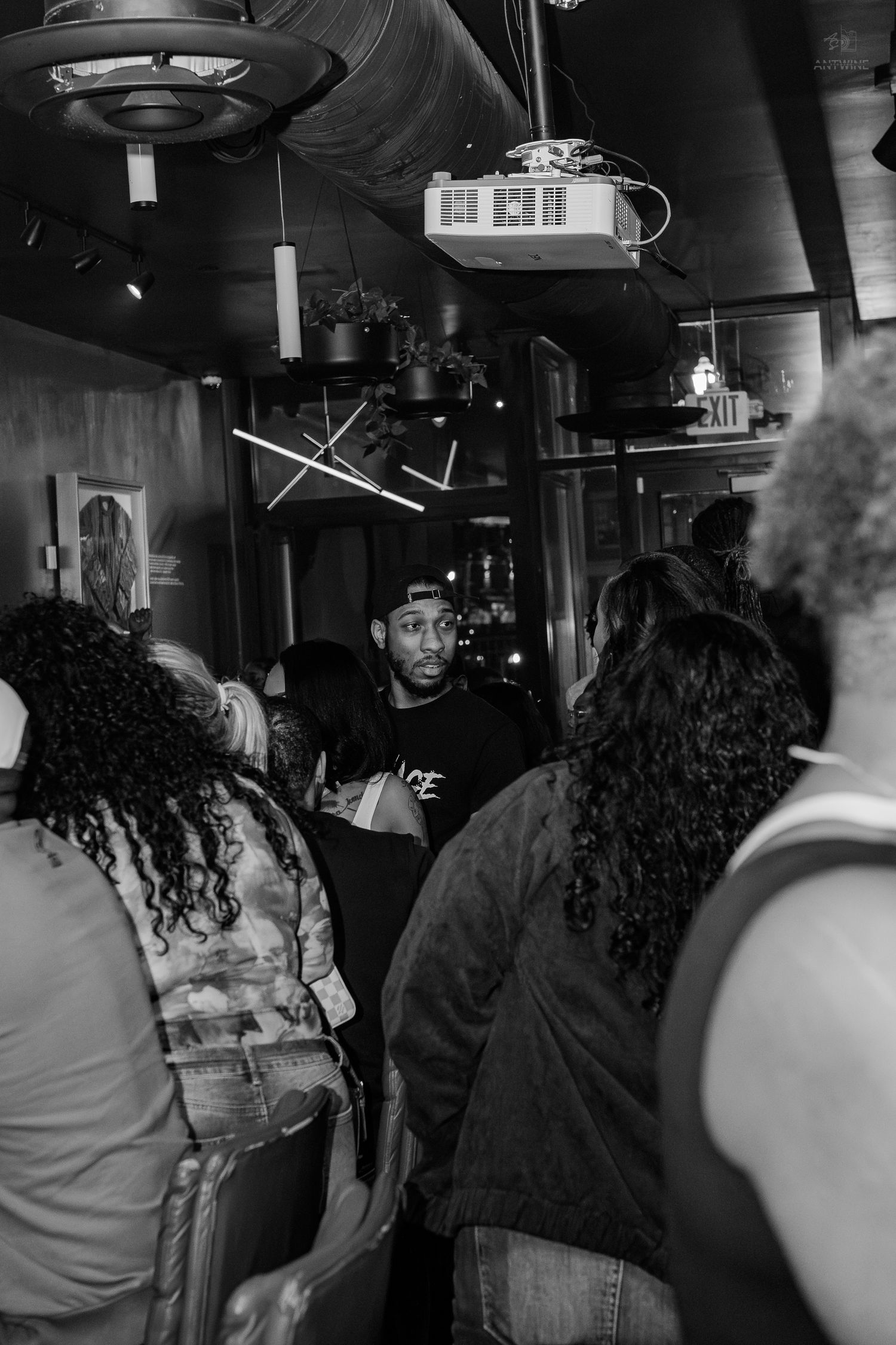 Black and white photo of a crowded indoor bar with people socializing under dim lights and a projector.