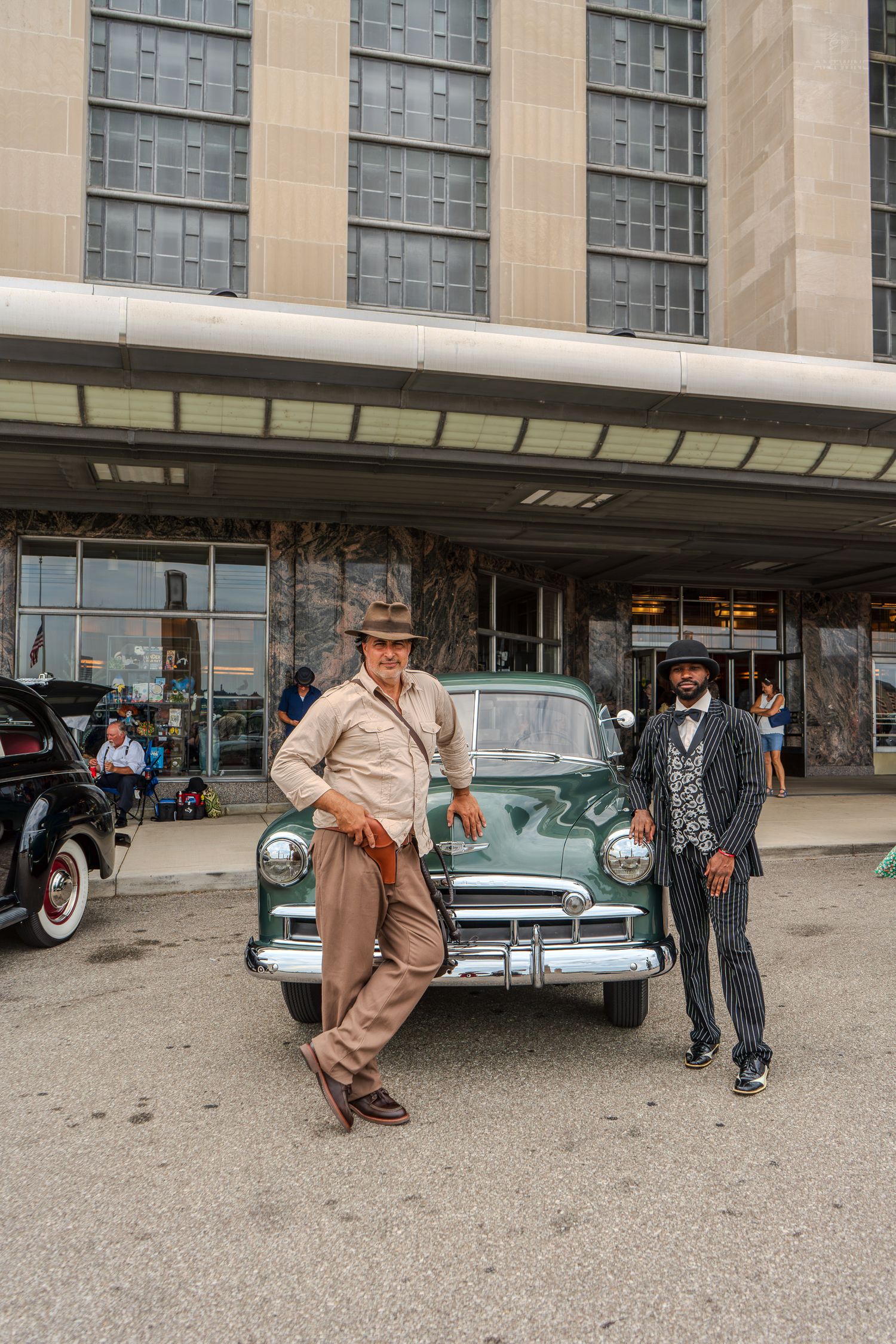 Two men in vintage attire pose beside a classic green car outside a retro building.