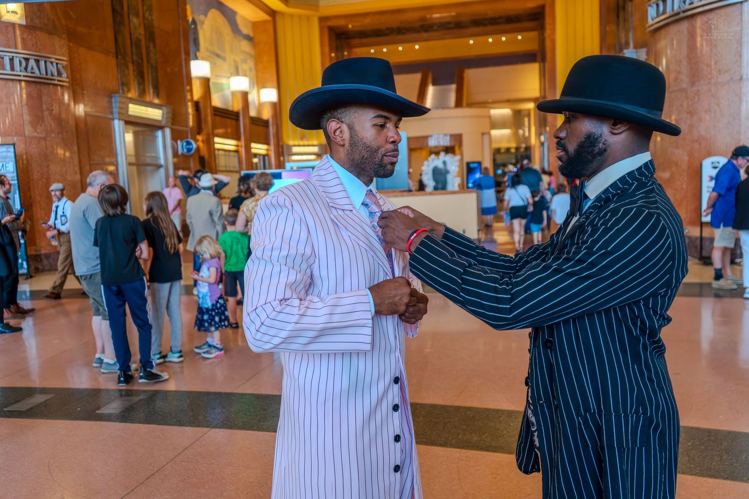 Two well-dressed Black men in pinstripe suits and wide-brim hats adjusting ties in a grand train station.