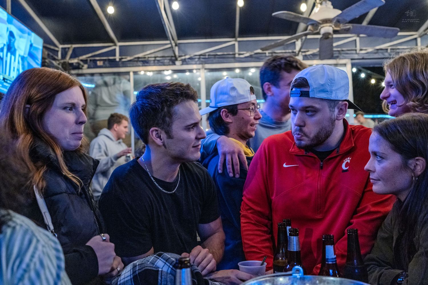 Group of young adults gathered at an outdoor bar venue, engaged in conversation under string lights at night.