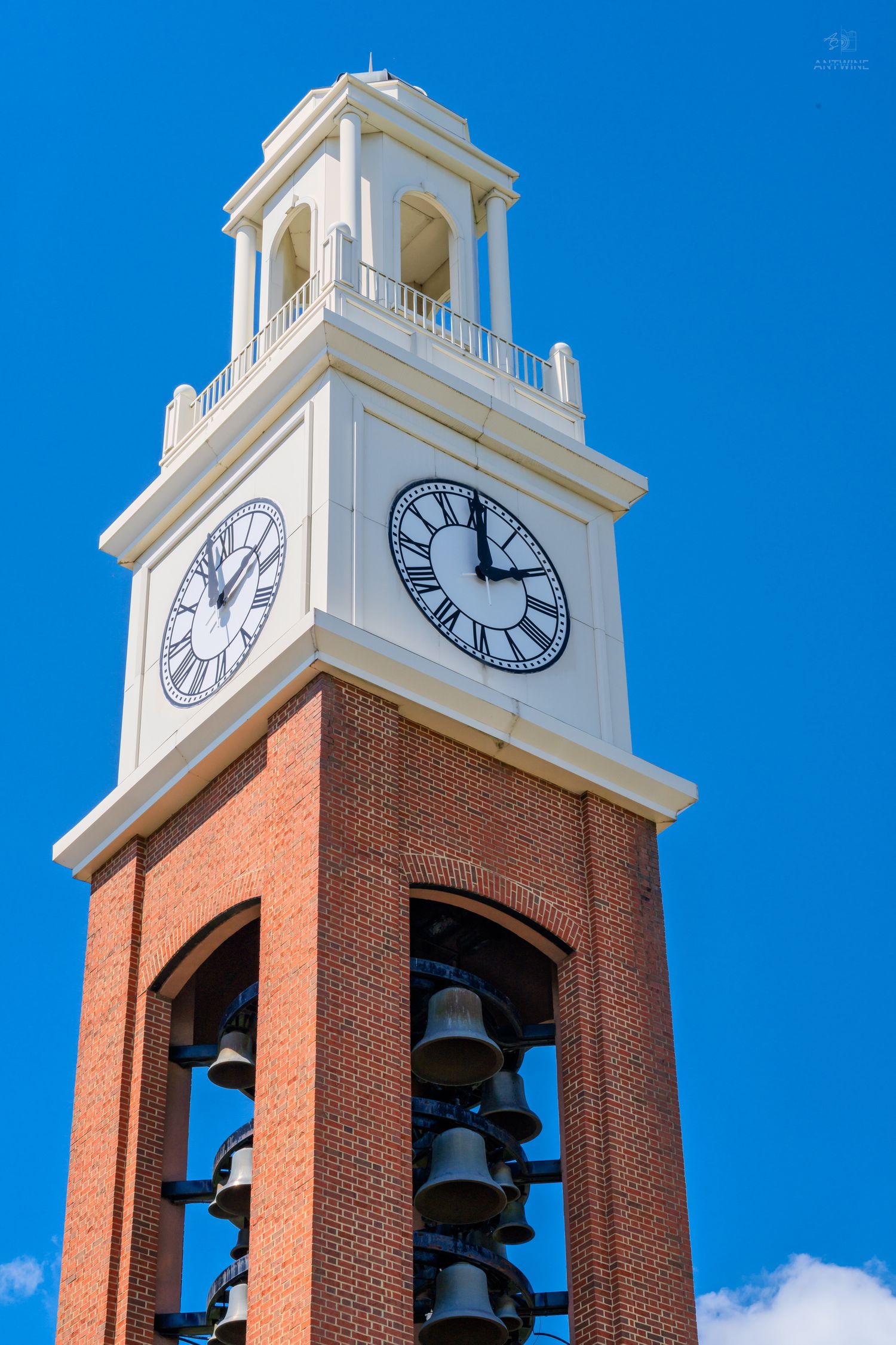 Red brick clock tower with bells and white cupola against a clear blue sky, showing nearly 12 o'clock on Roman numeral clock faces.