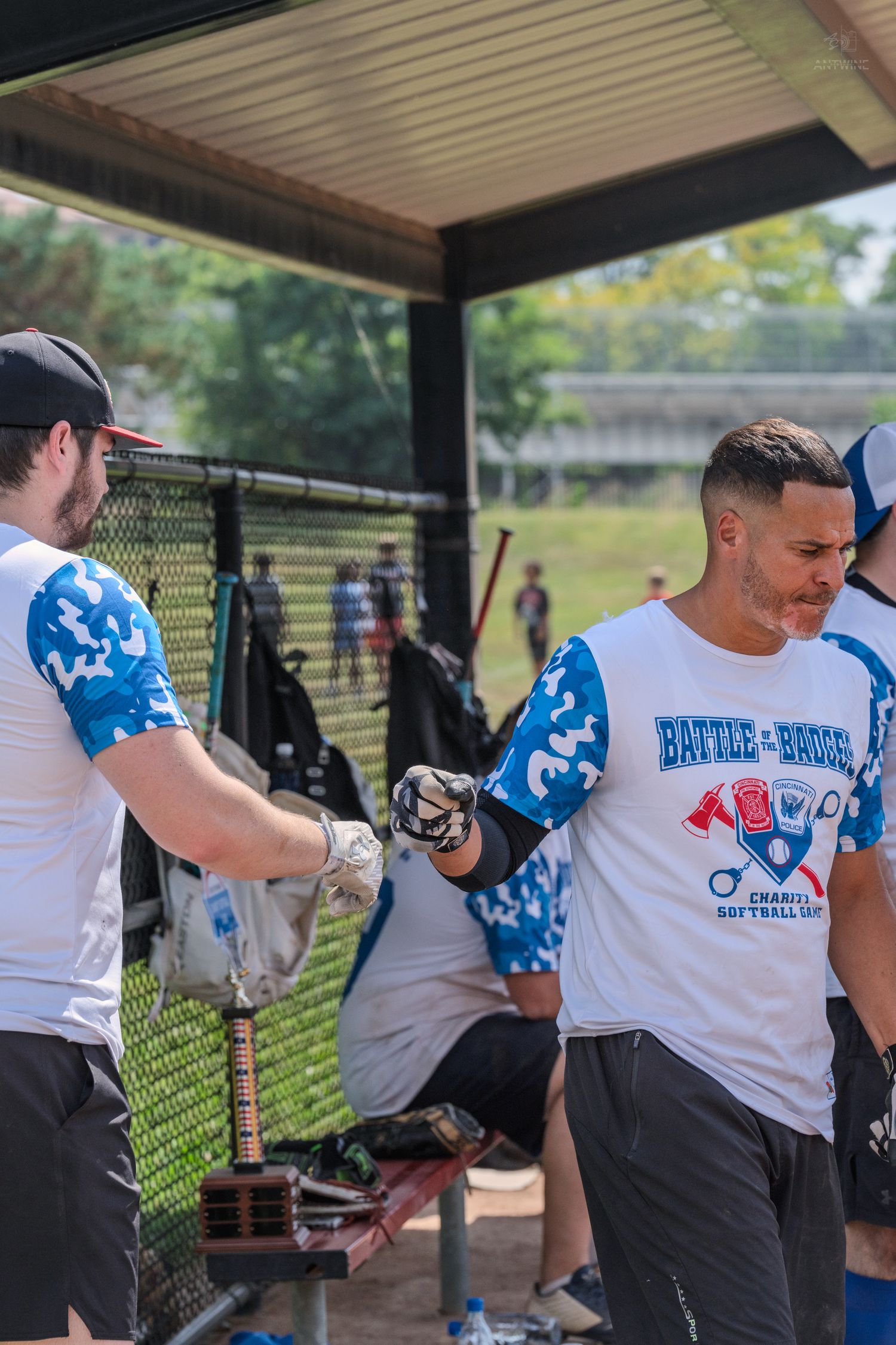 Two players in Battle of the Badges charity softball uniforms exchange a fist bump near the dugout.