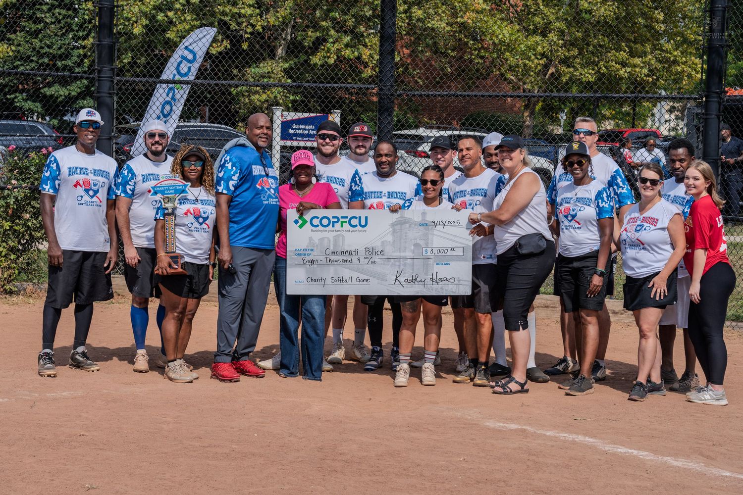 Group of people holding large COPFCU charity check at outdoor softball field event.