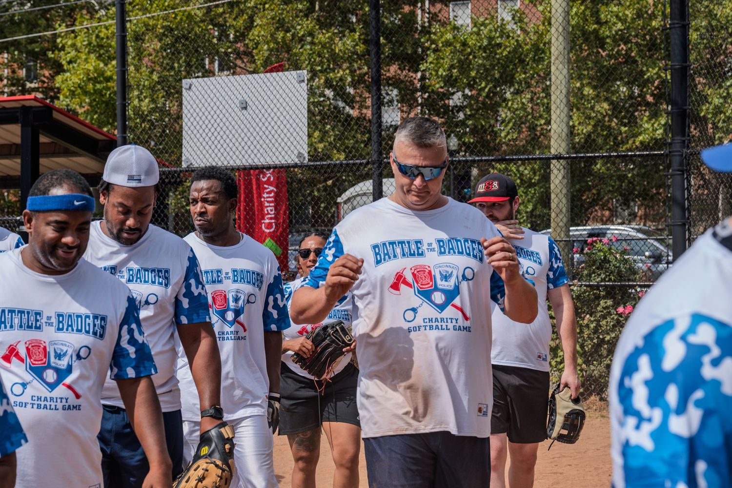 Players in Battle of the Badges softball uniforms walk together on a sunny outdoor baseball field.