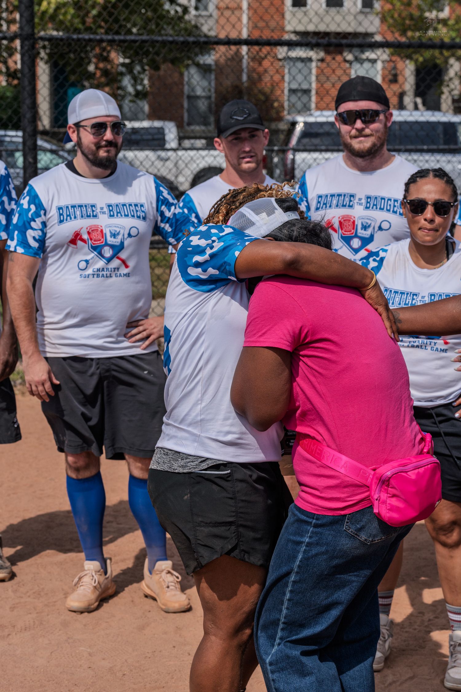 People in Battle of the Badges charity softball event shirts hugging on a baseball diamond on a sunny day.