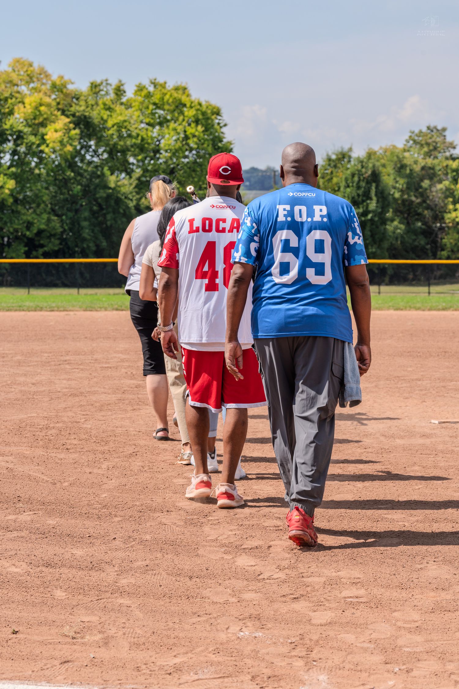 Players in softball jerseys walking across a dirt baseball field on a bright sunny day.