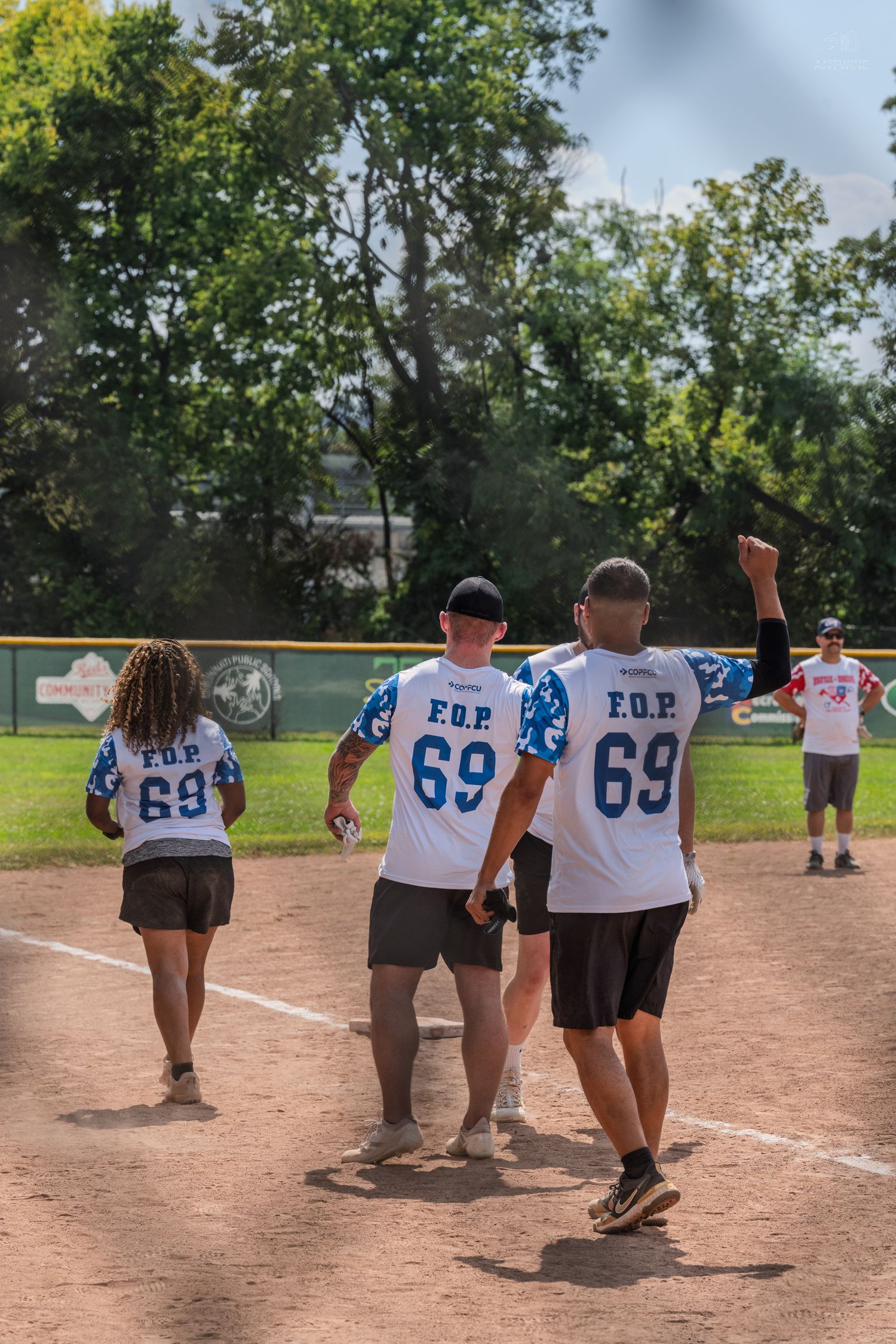 FOP softball team players wearing number 69 jerseys celebrate on baseball diamond during sunny outdoor game.
