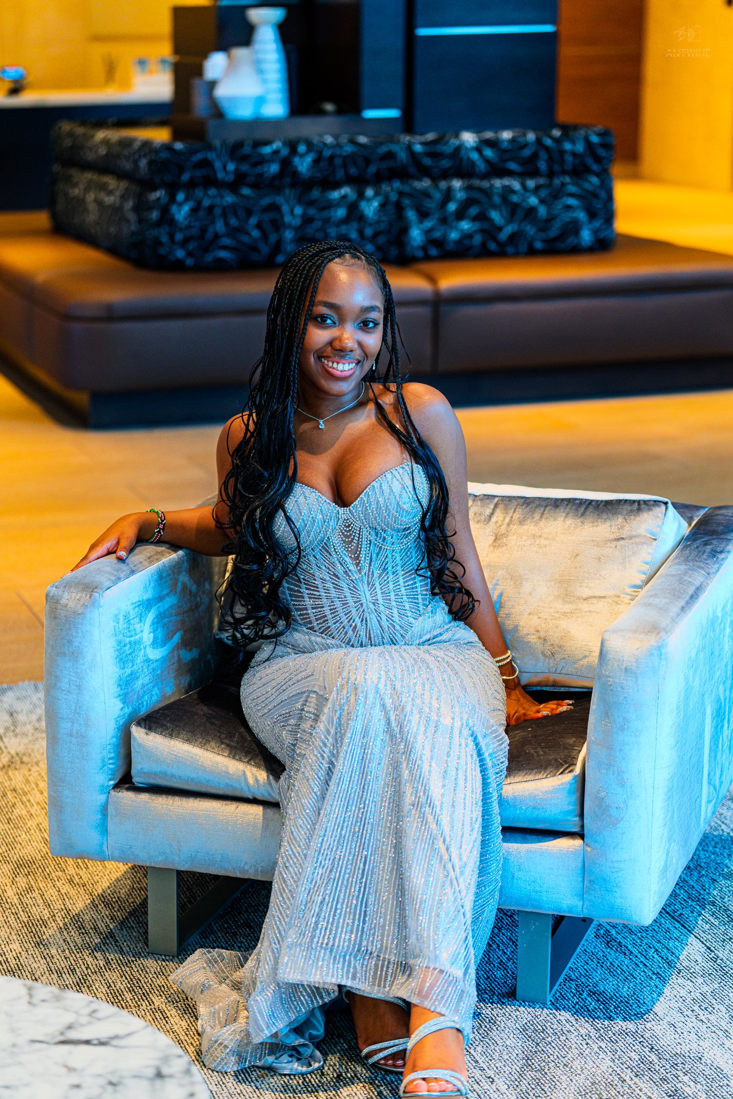 A smiling woman in a silver beaded gown sits elegantly in a velvet chair in a modern upscale venue.