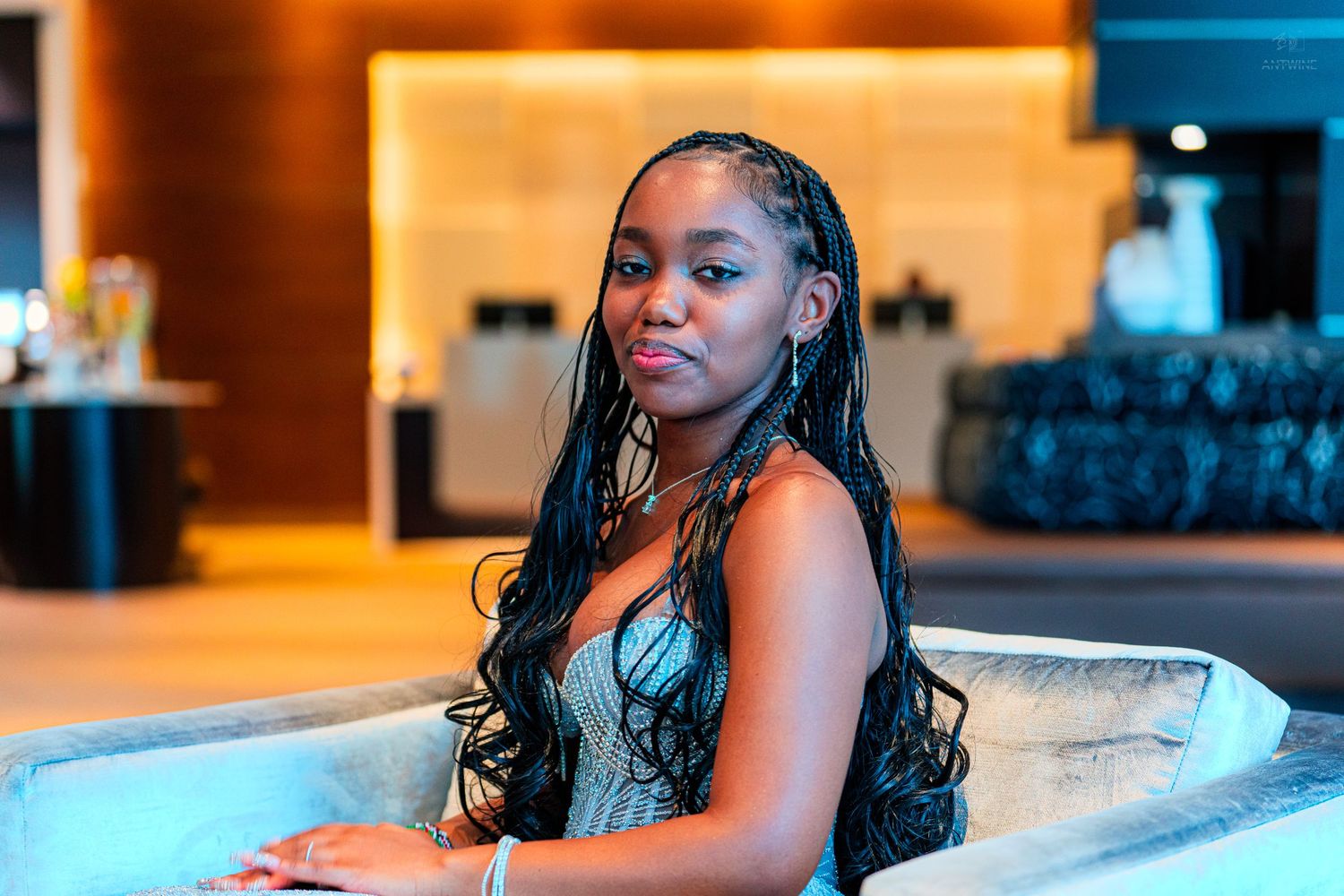 Young woman with long braided hair poses elegantly in a modern hotel lobby, wearing a stylish outfit.