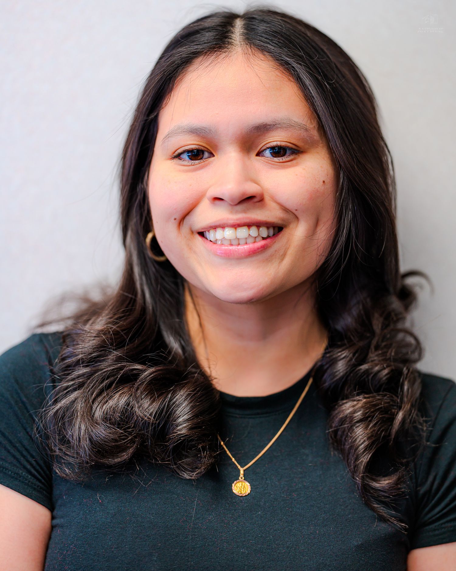 Young woman with long dark curly hair smiling, wearing a dark green top and gold necklace.
