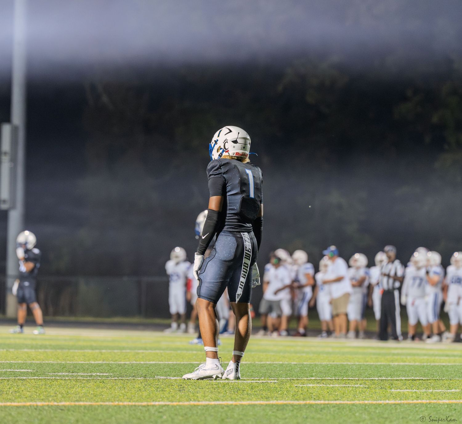 High school football player in black uniform stands on field under bright stadium lights during a game.