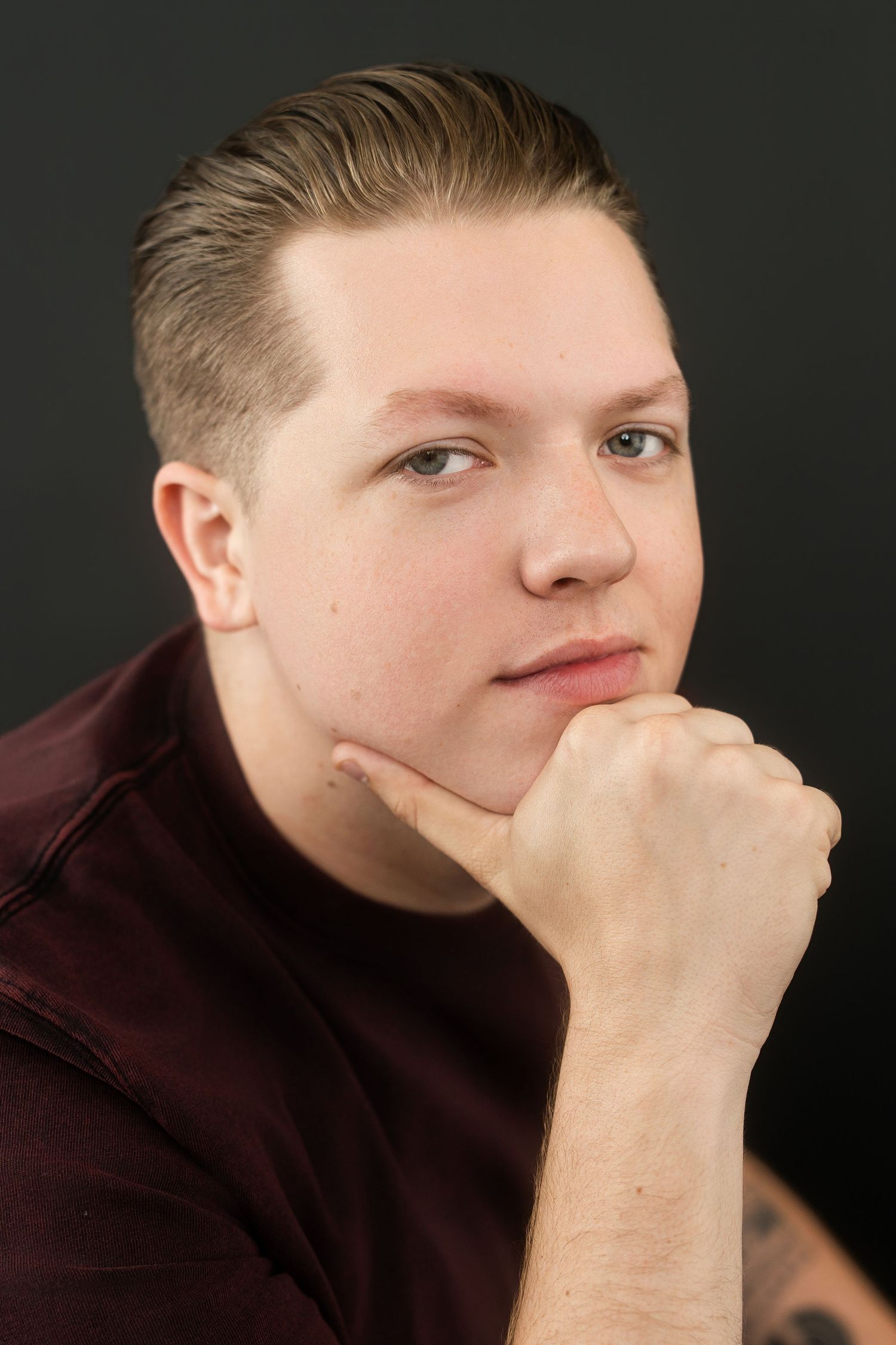 Young man in maroon t-shirt resting chin on fist, thoughtful pose against dark background.