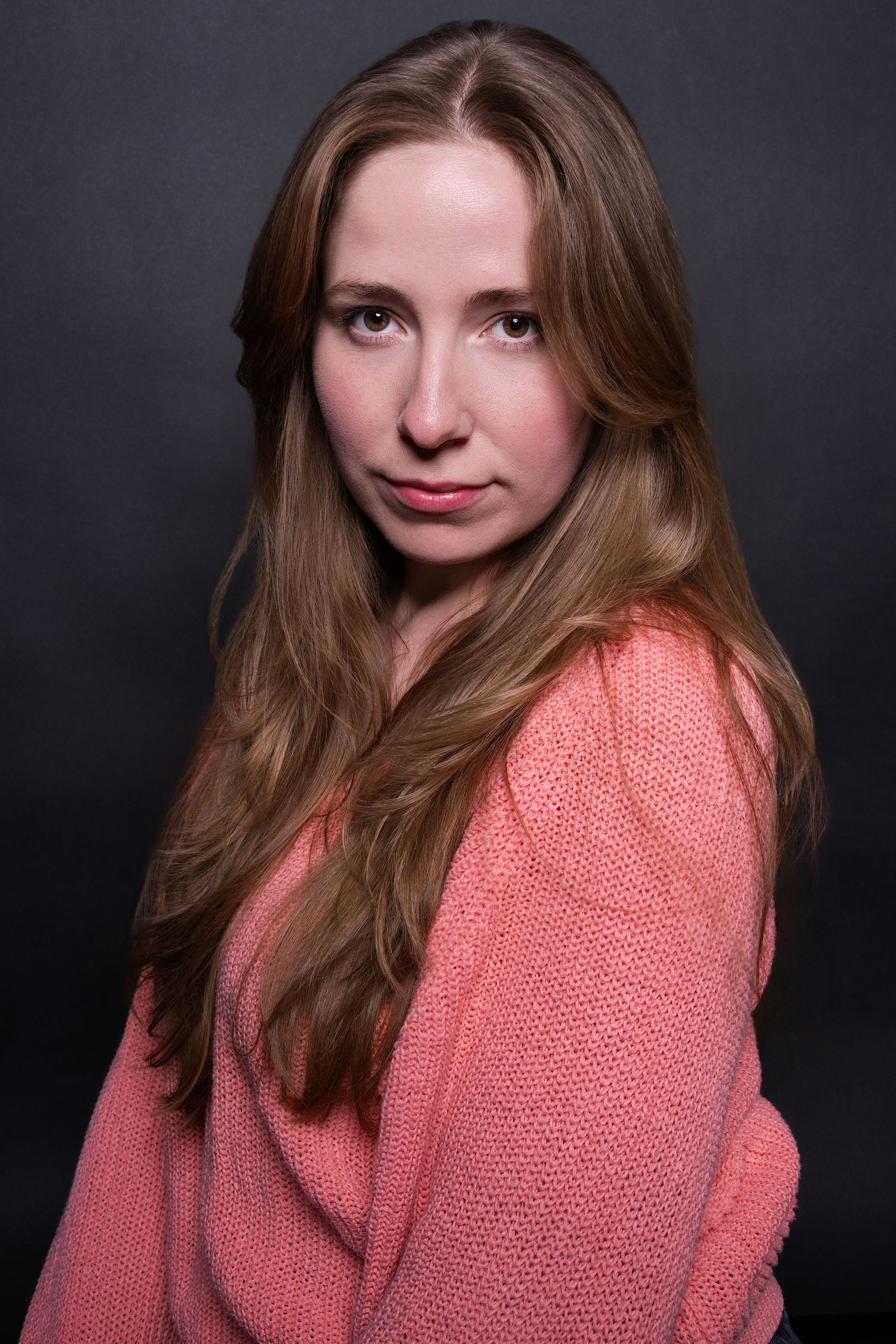 Woman with long auburn hair in a pink knit sweater gazing at camera against a dark background.