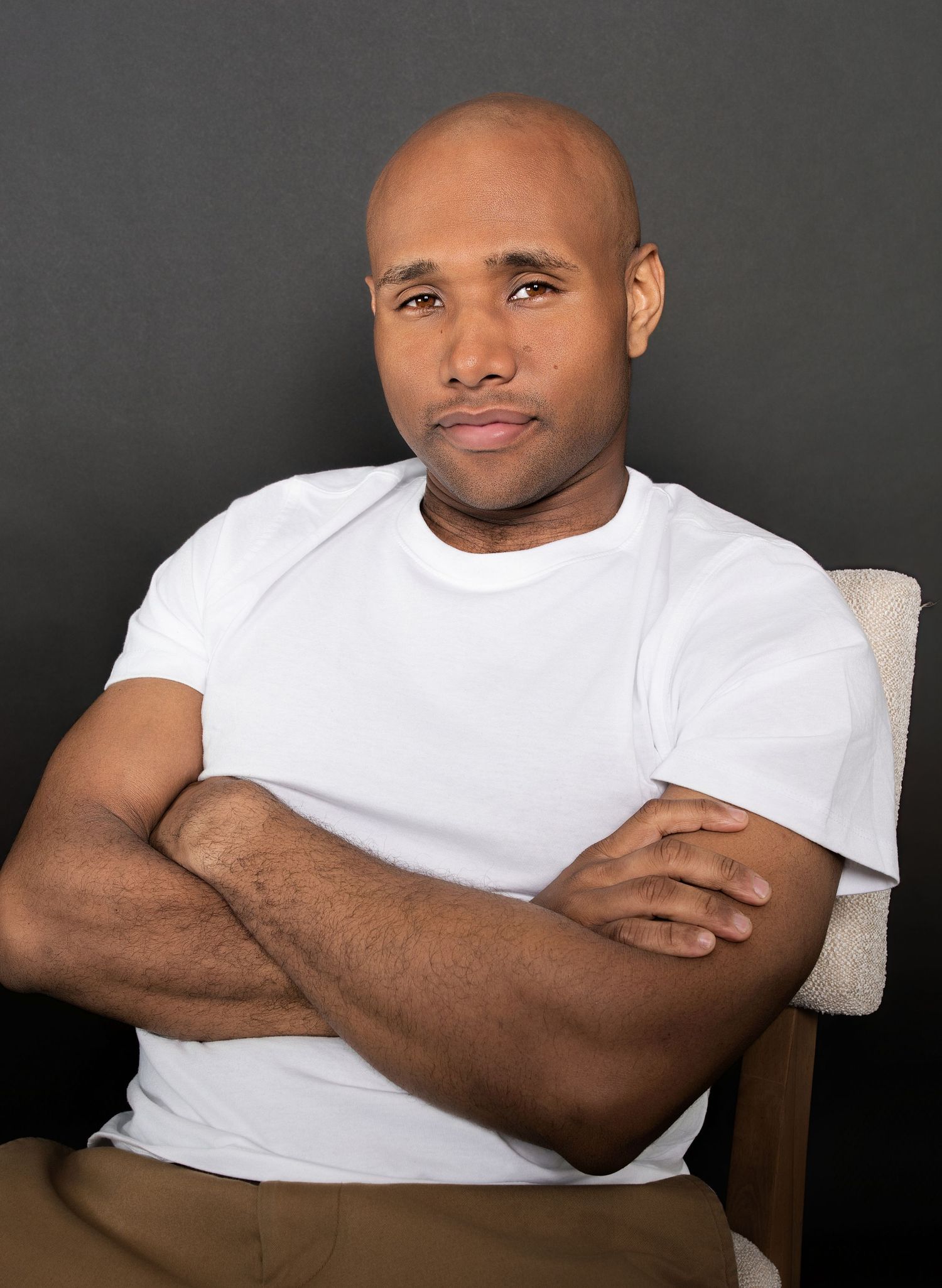 Bald man in white t-shirt sitting with arms crossed in chair against dark background.