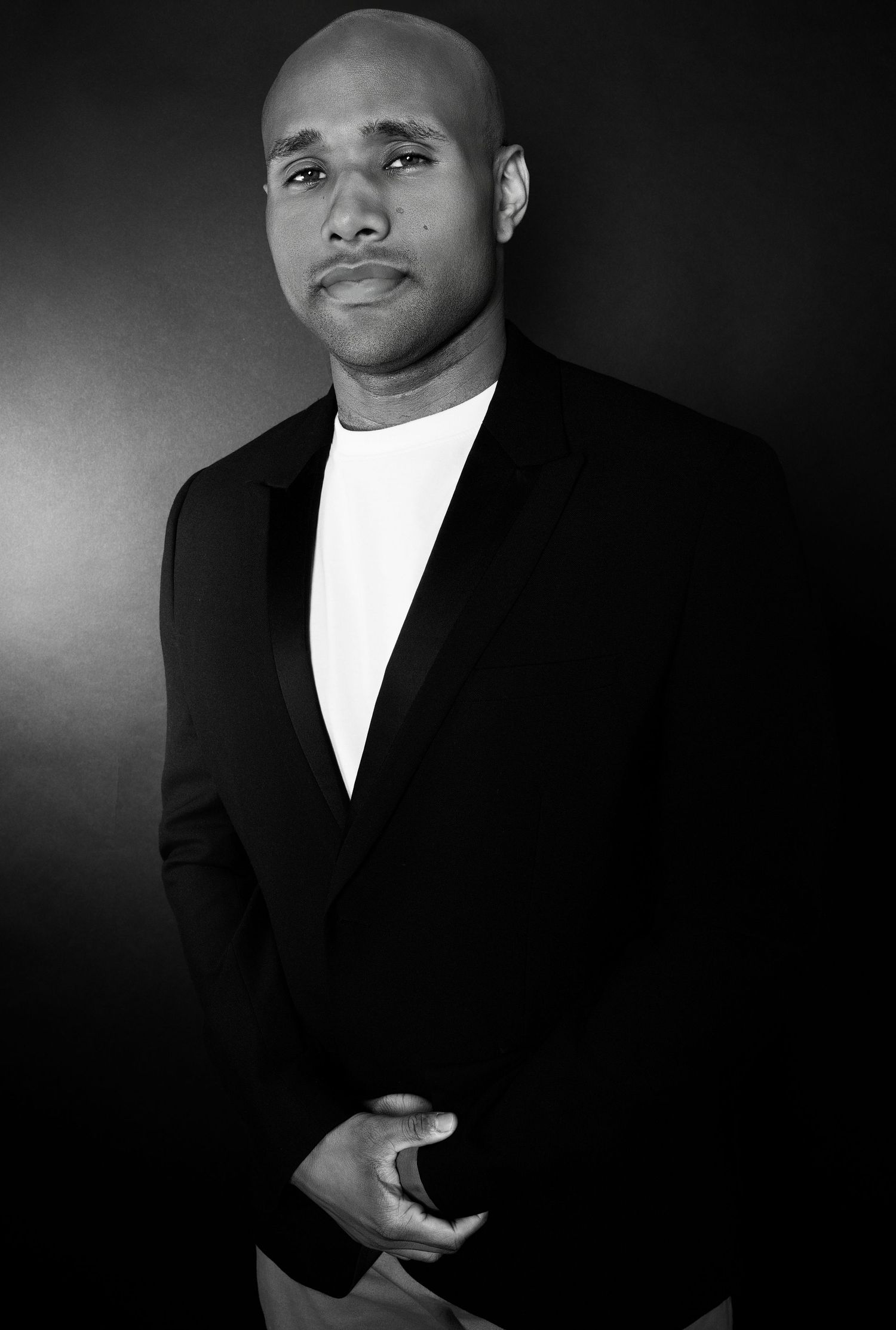 Bald Black man in black blazer and white shirt posing in dramatic black and white portrait.