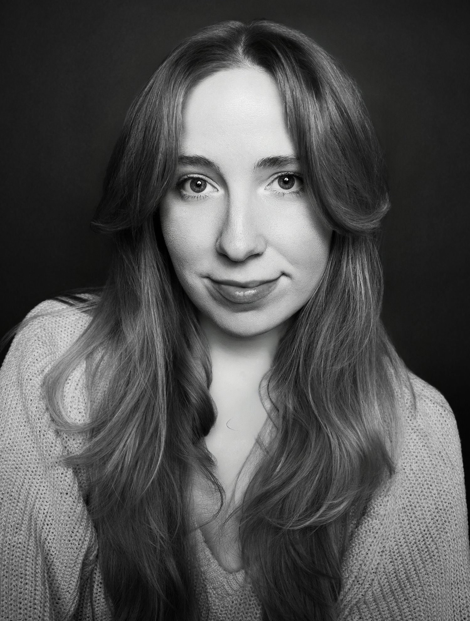 Black and white headshot of young woman with long wavy hair in knit sweater against dark background.
