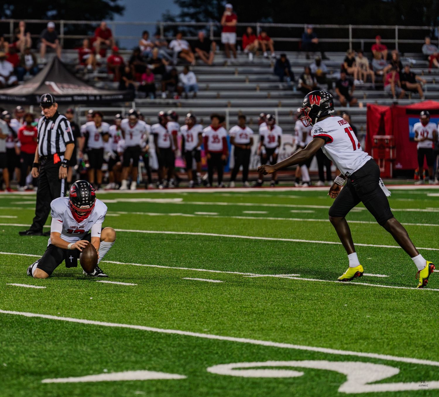 A football player prepares to kick a field goal while a teammate holds the ball on a green field.