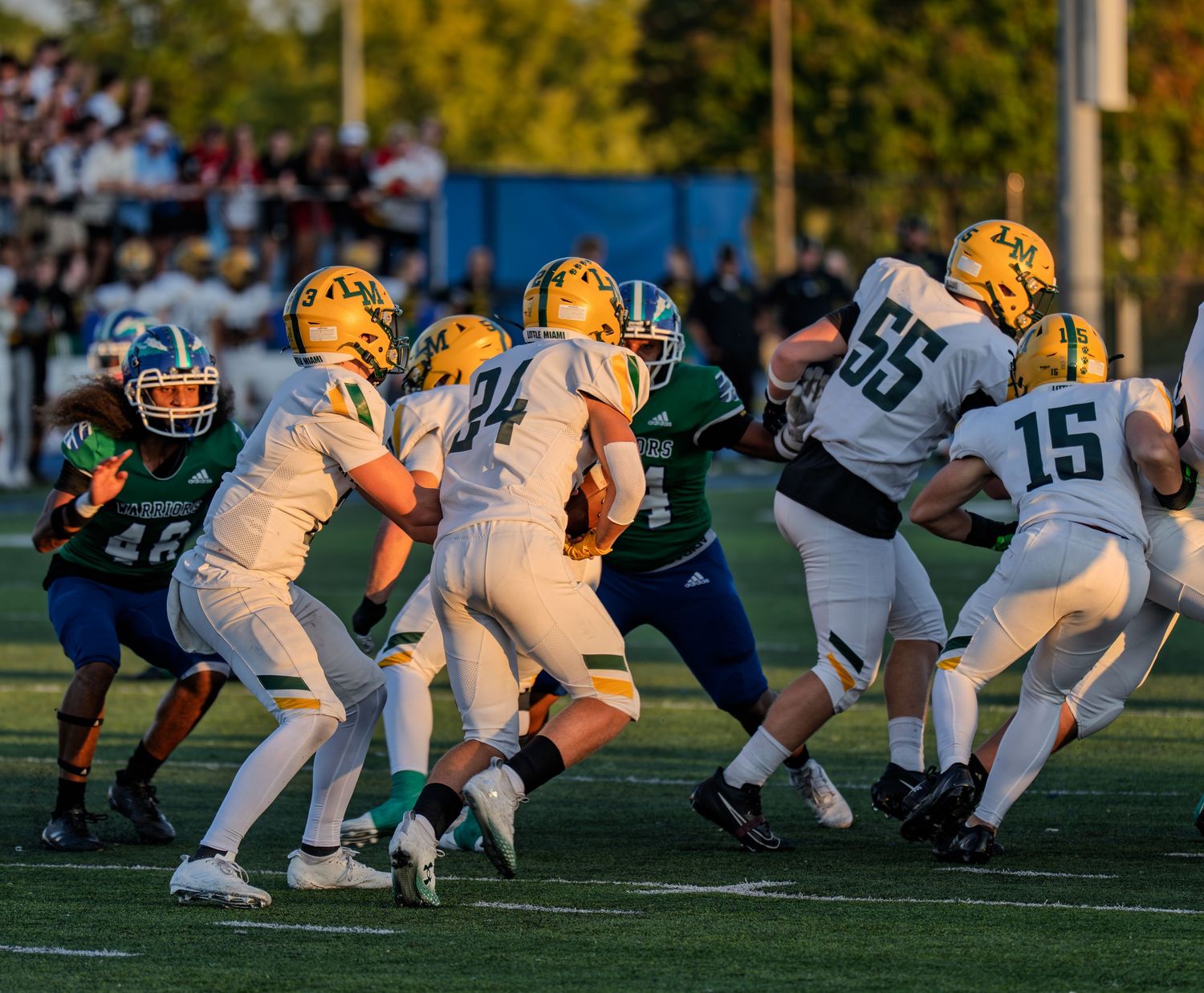 Football players in white and gold uniforms execute a running play against green-uniformed defenders in a high school game.