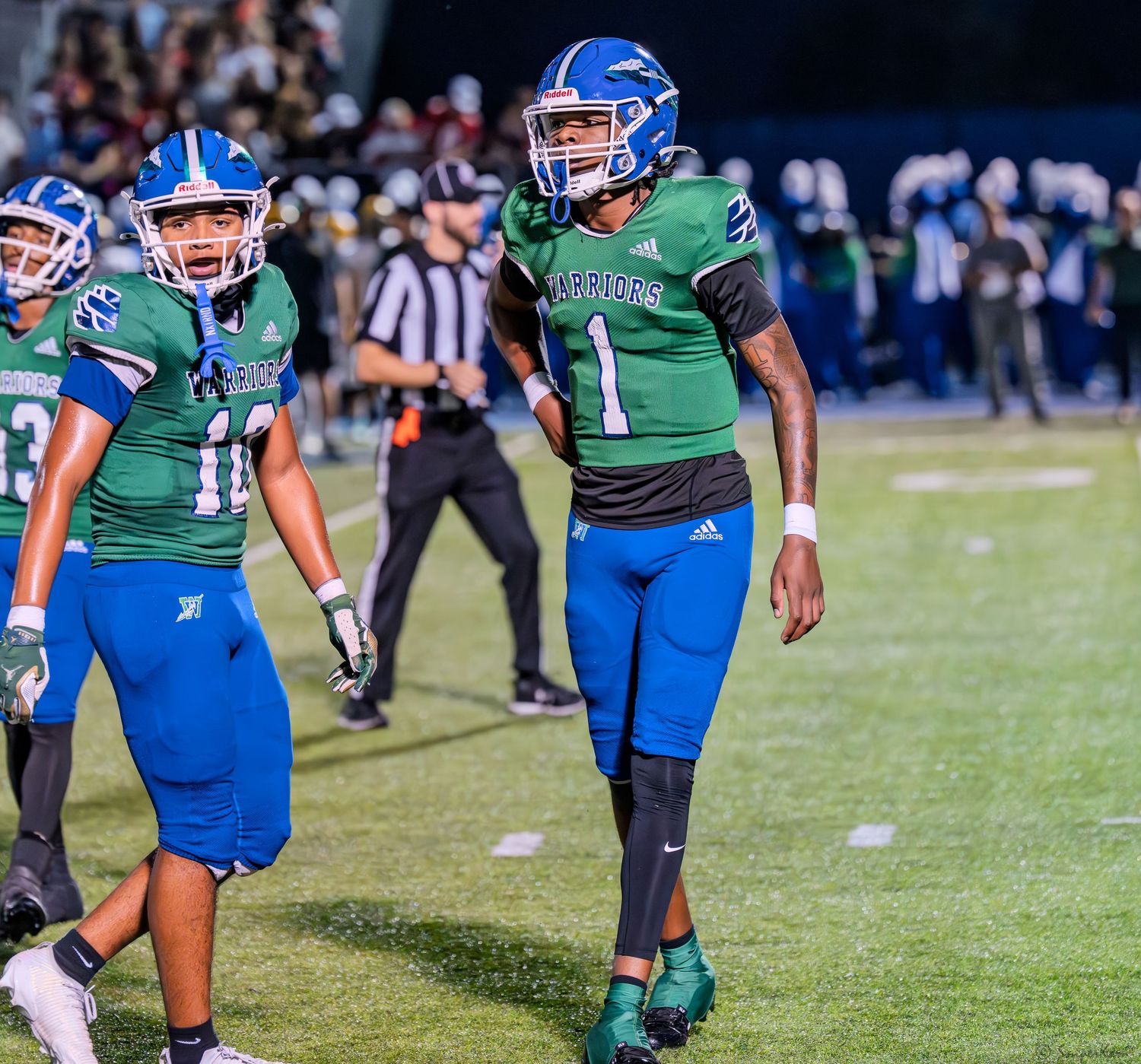 Three high school football players in green and blue uniforms walk on the field during a night game.