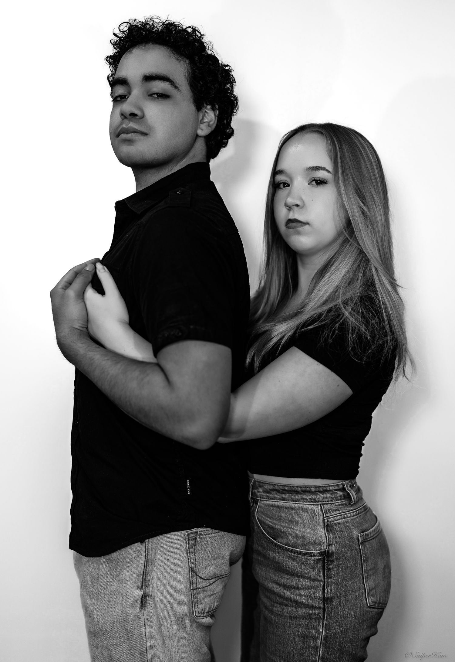 Black and white portrait of a young man and woman posing back-to-back against a clean white background.