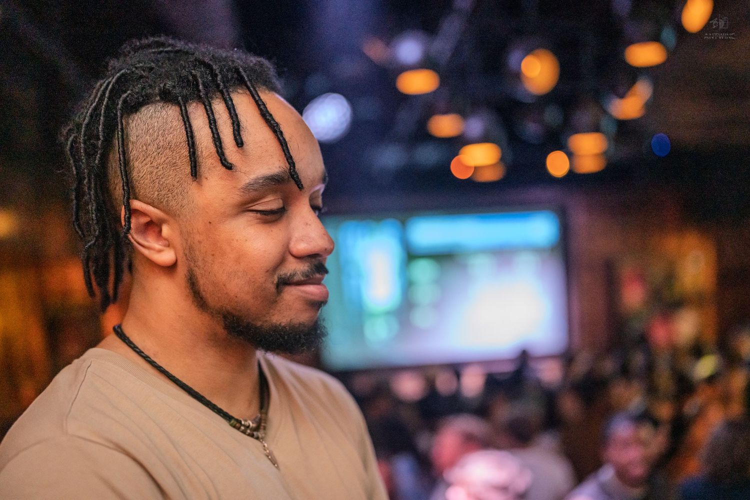 A young man with braided hair smiles softly in a dimly lit bar with bokeh lights behind him.