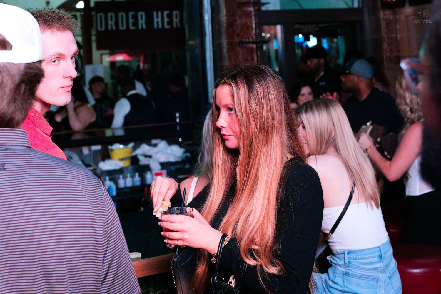 Young woman with long hair holding drink at busy bar event.