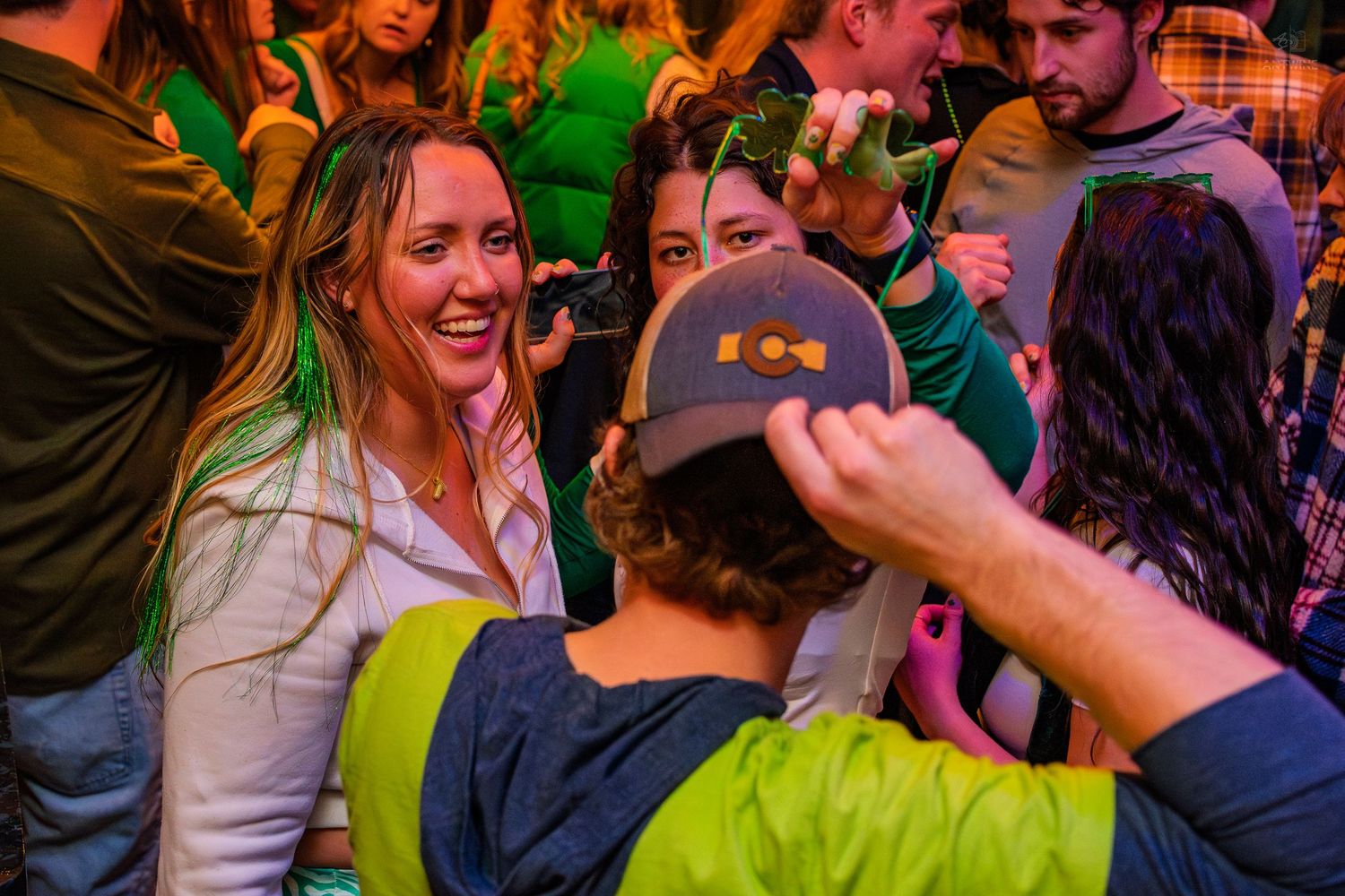 Smiling woman with green hair streaks enjoying a lively St. Patrick's Day bar celebration with friends.