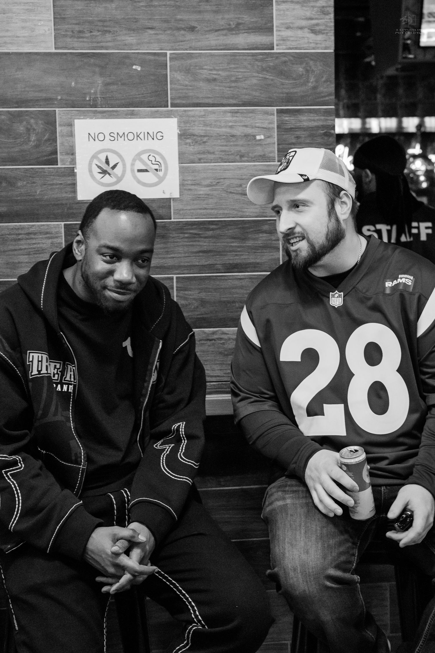 Black and white photo of two men sitting and chatting near a no smoking sign at a venue.