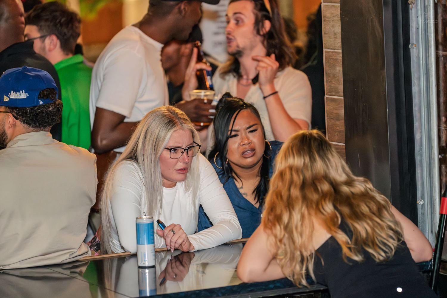 Group of young women having an animated conversation at a crowded bar or nightclub setting.