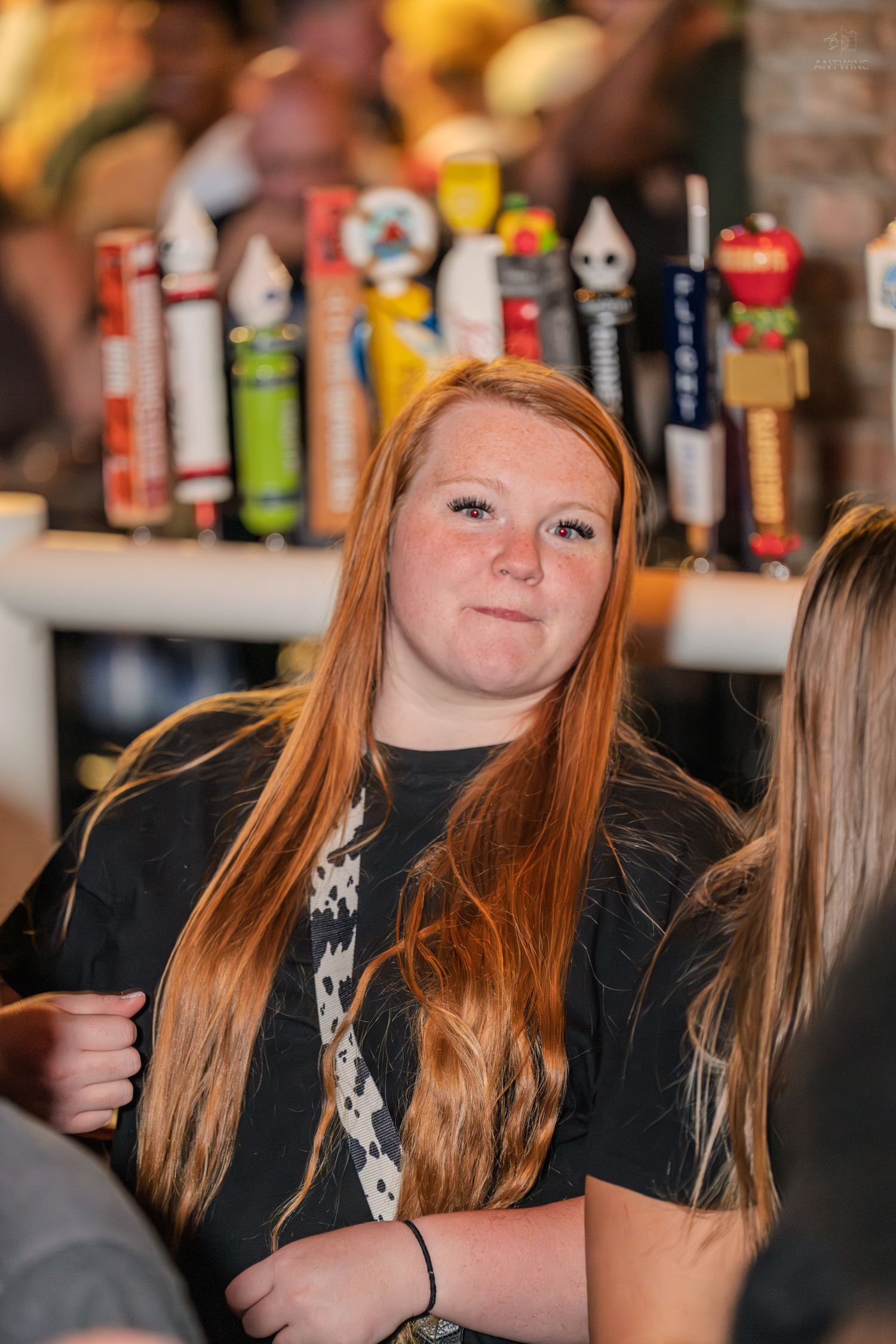 Young redheaded woman in a black t-shirt smiling at a bar with colorful beer taps behind her.