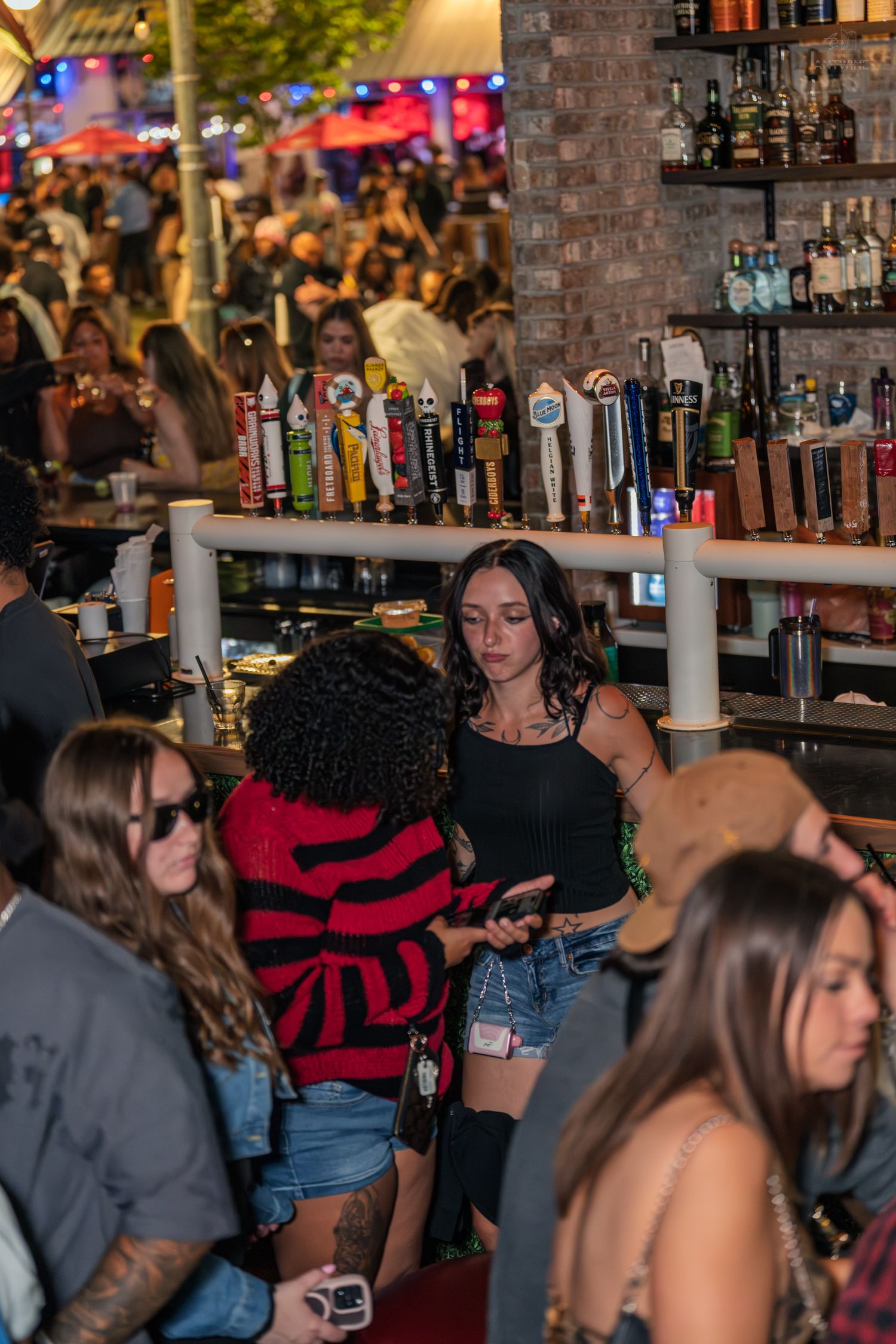 Young woman in black top checking phone near bar with multiple draft beer taps at nightclub.