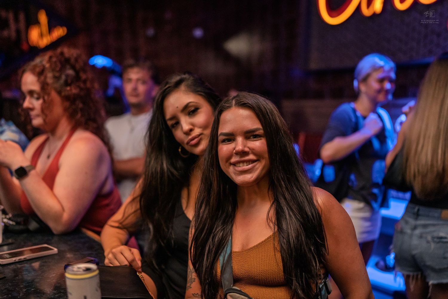 Two dark-haired women smiling at the camera inside a neon-lit bar.