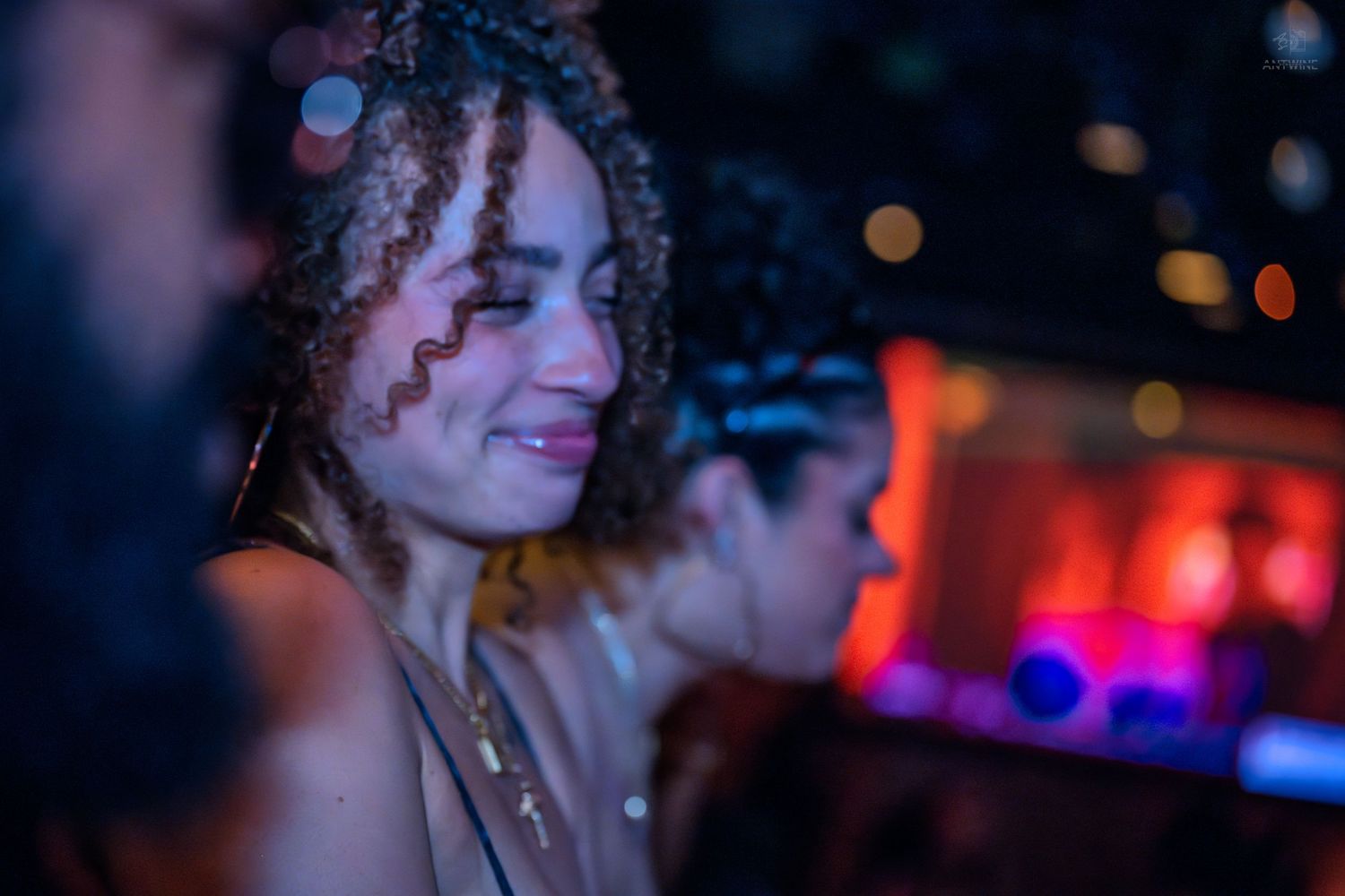 Young woman with curly hair dancing and smiling at a nightclub with red stage lights behind her.
