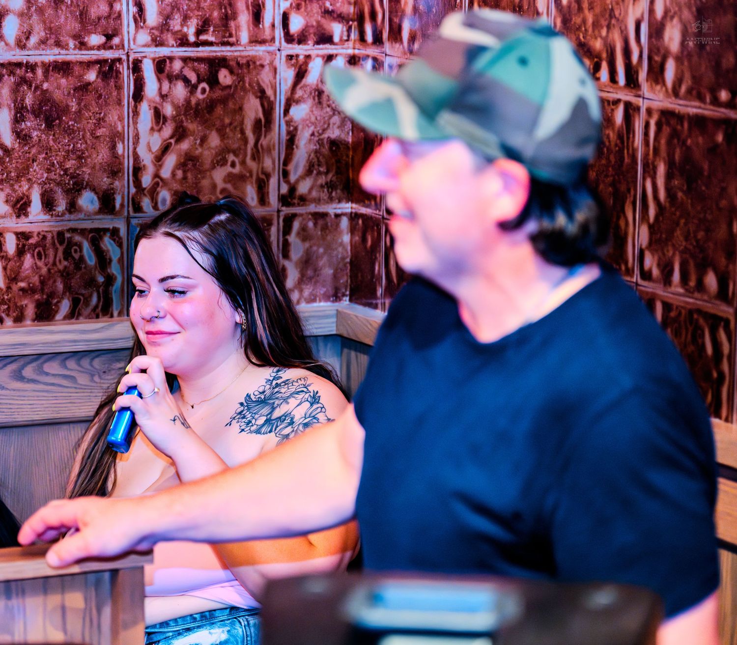 Woman with shoulder tattoo holding microphone beside man in camo cap at bar.