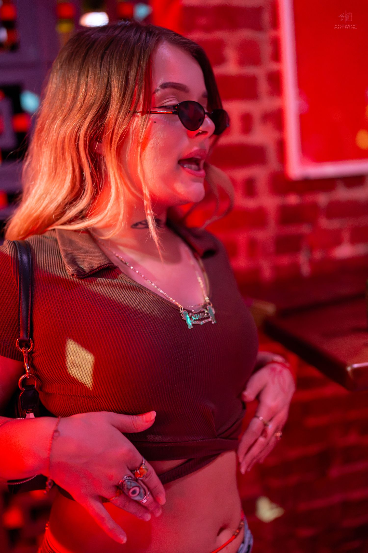 Woman in sunglasses and crop top poses under red neon lights at a bar, wearing statement rings and a chain necklace.