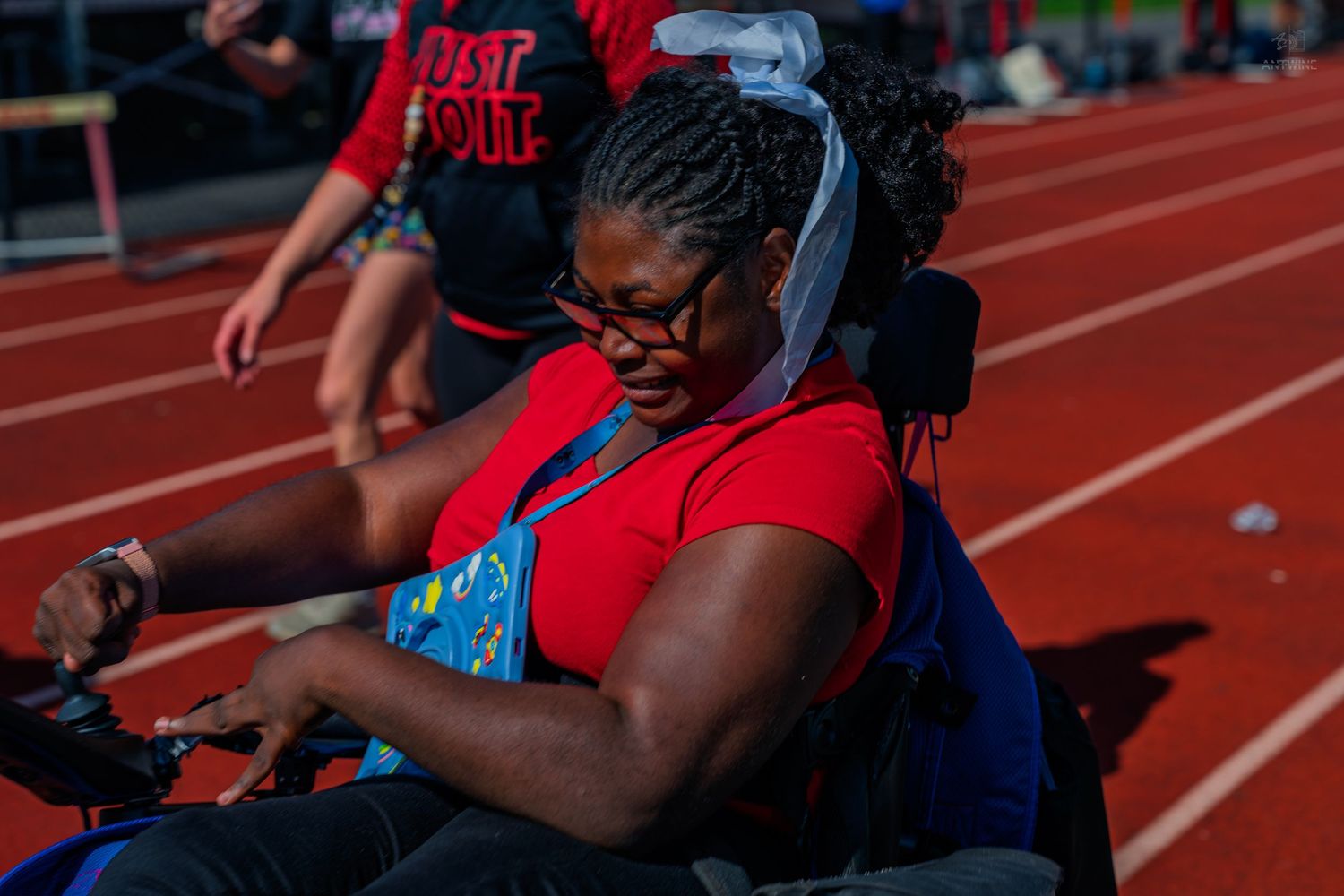 A young Black woman in a red shirt sits in a wheelchair on a red track during an outdoor athletic event.