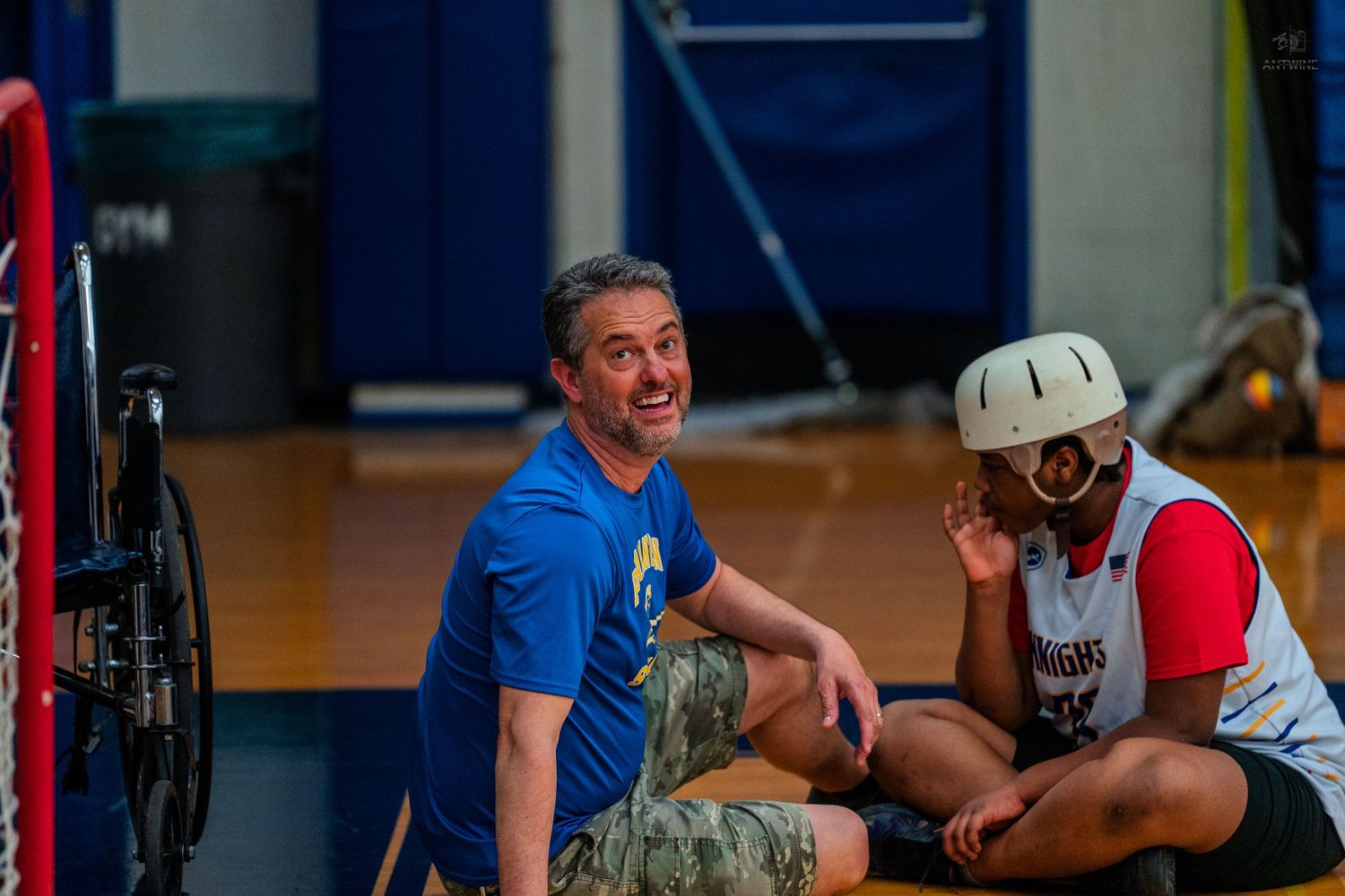 Two men sit on a gym floor laughing, one wearing a blue shirt and the other a helmet.