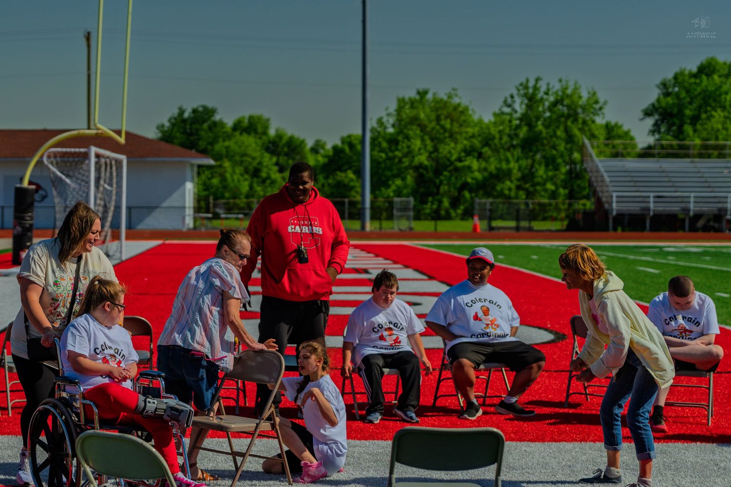 Group of people in white shirts gathered on a red track at an outdoor sports field on a sunny day.