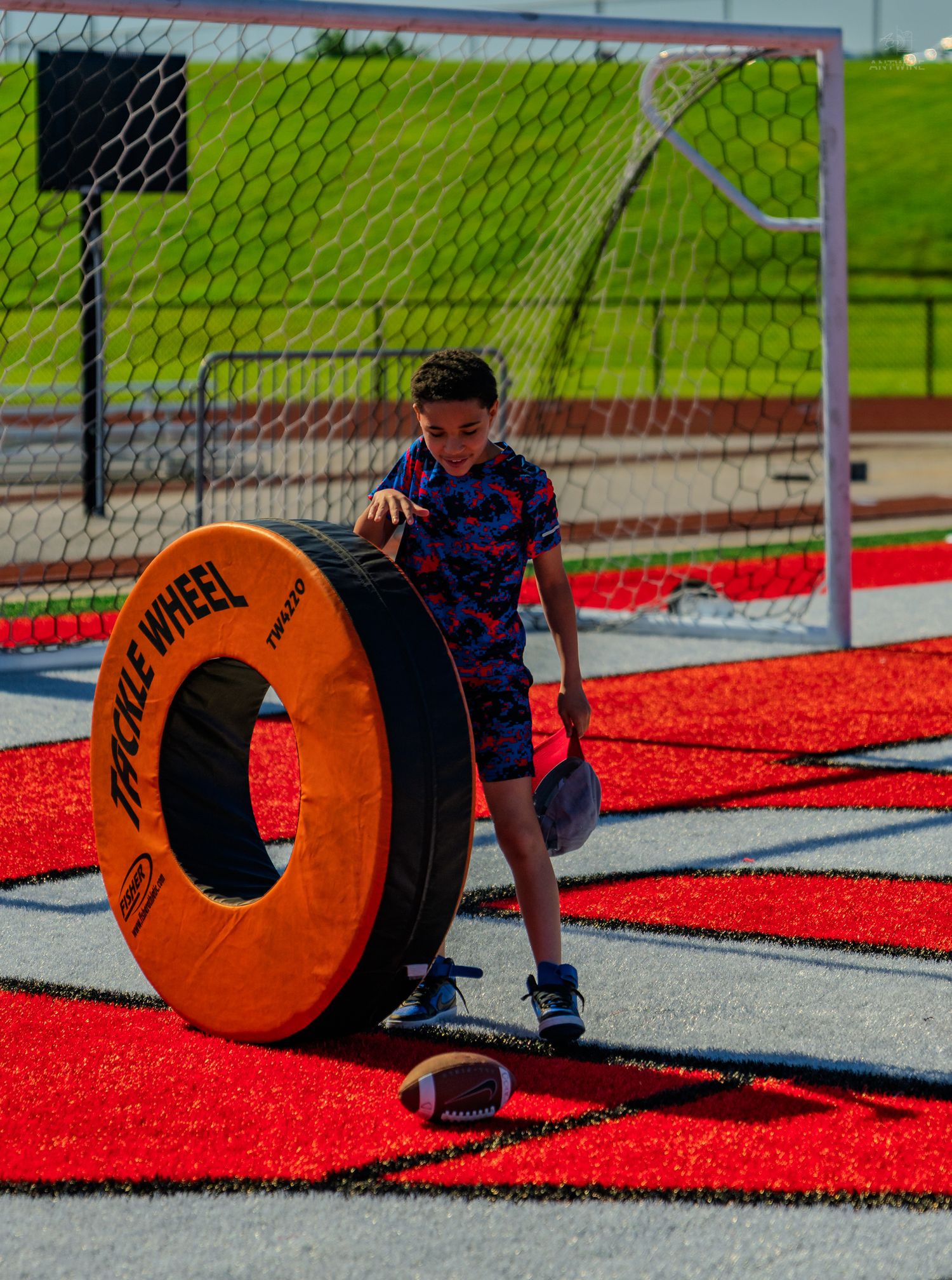 A young boy pushes an orange Tackle Wheel training tire on a red turf field near a soccer goal.