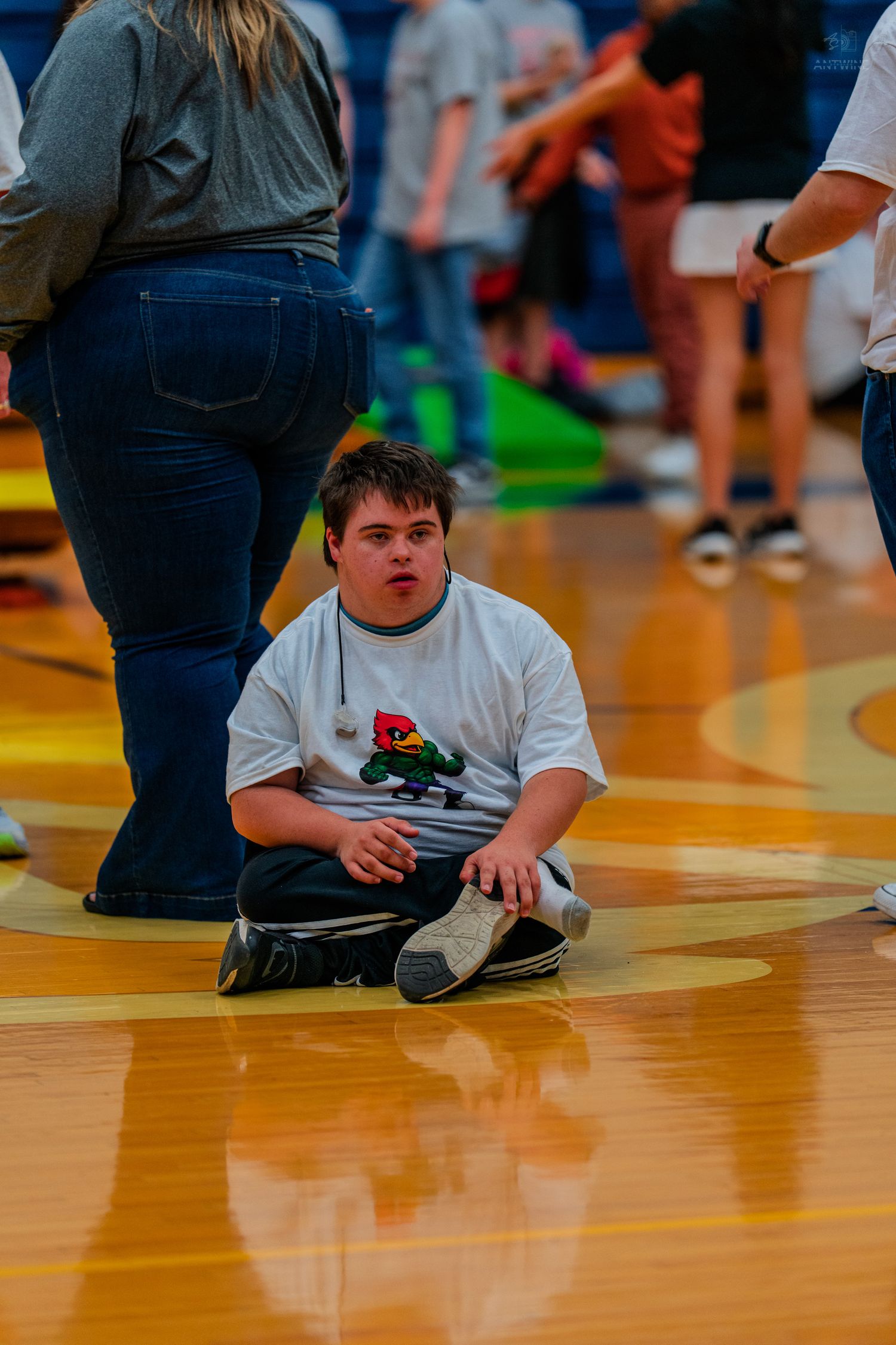 Young person with Down syndrome sitting on gymnasium floor during a school sports event.