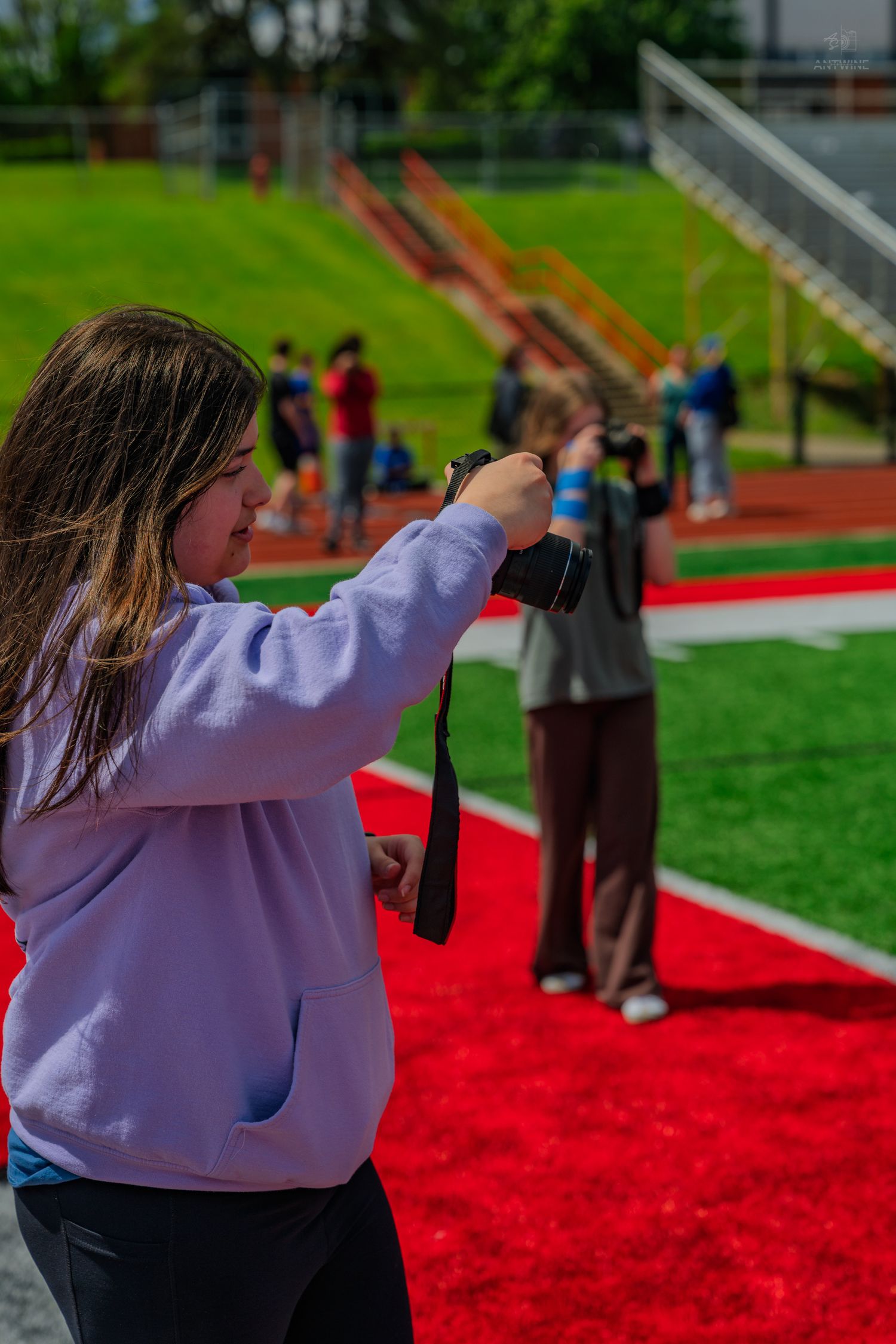 Girl in lavender hoodie photographing at a red turf sports field on a sunny day.