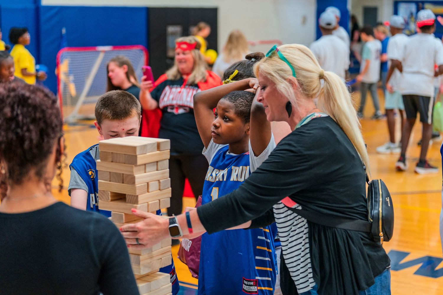 Children and a teacher play Jenga together at a school gym event, enjoying a fun indoor activity.