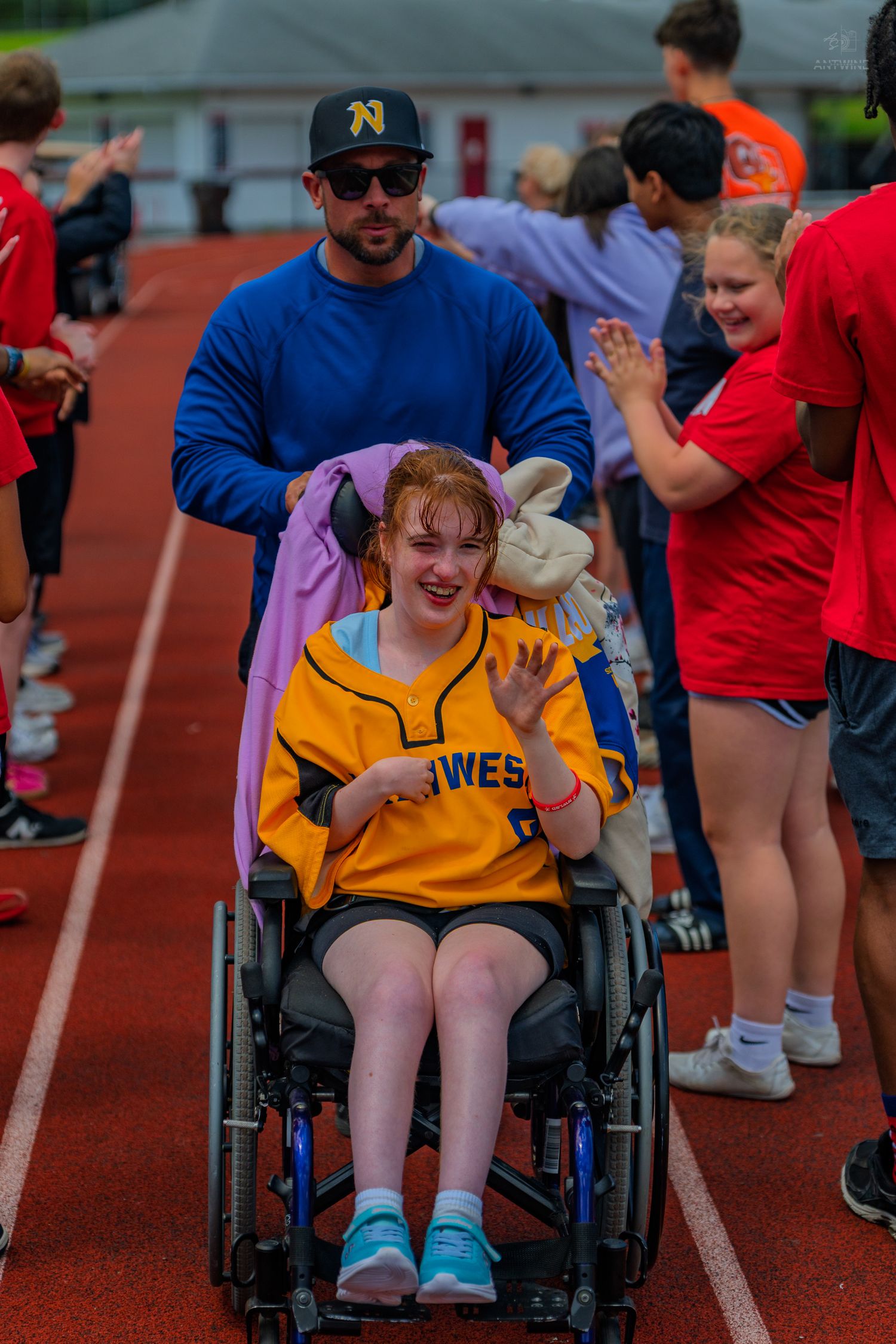 A smiling girl in a wheelchair wearing a yellow jersey is pushed along a track while people high-five her at a special sports event.